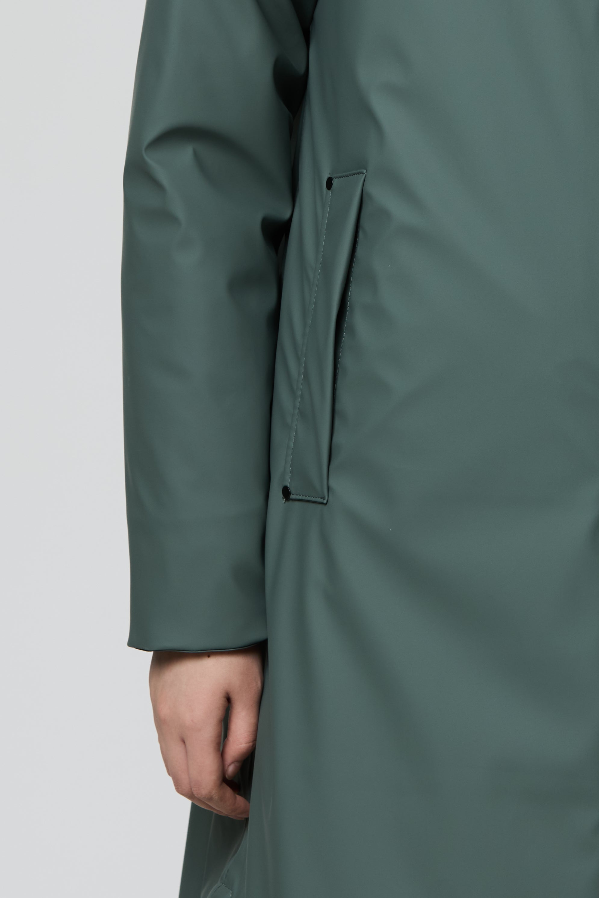 Close-up of a person wearing a long, dark green raincoat or trench coat, showing the sleeve, side pocket, and hand against a light gray studio background.