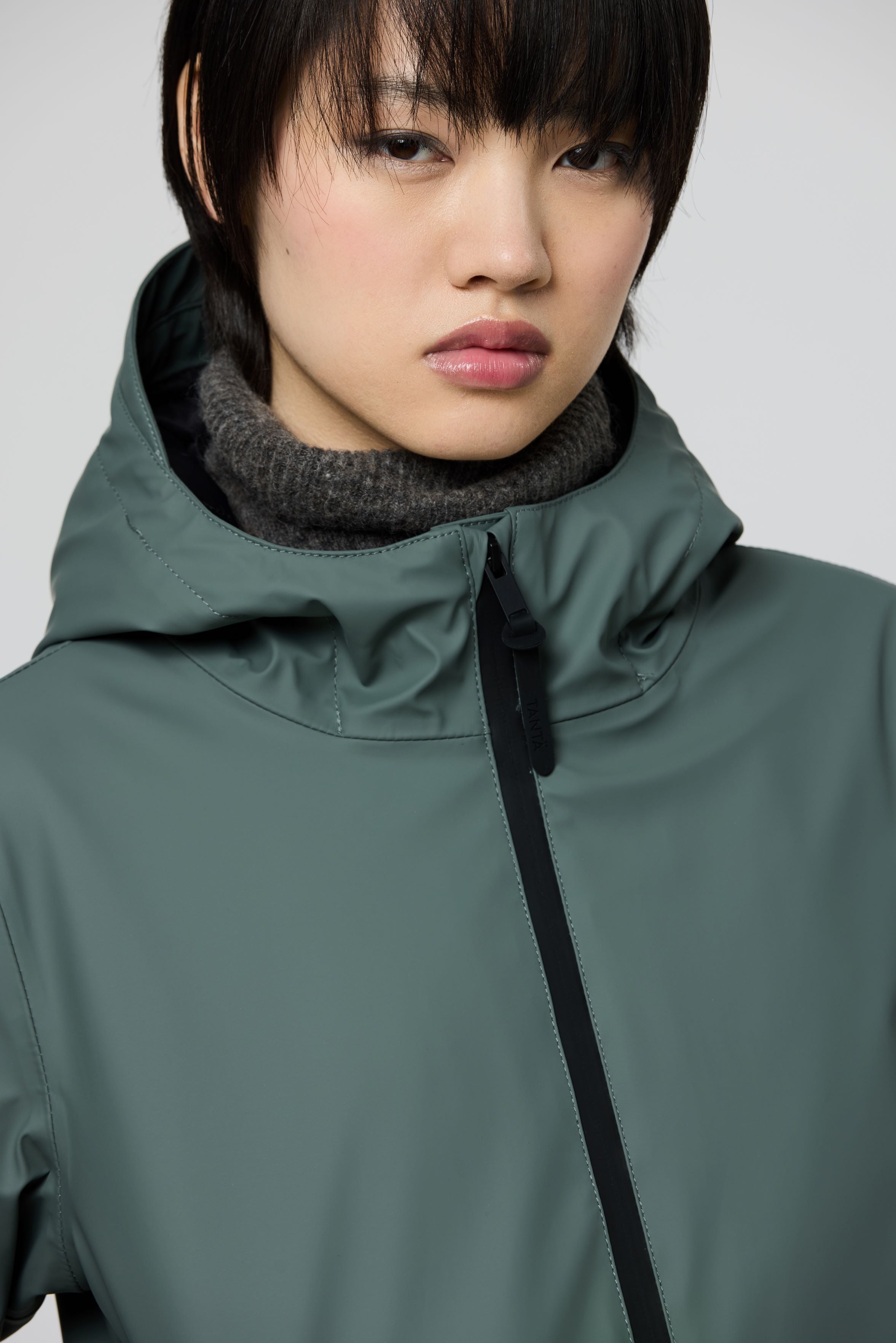 Close-up of a model wearing a muted sage green hooded jacket with a black zipper and a dark gray knit neck warmer against a light gray background.