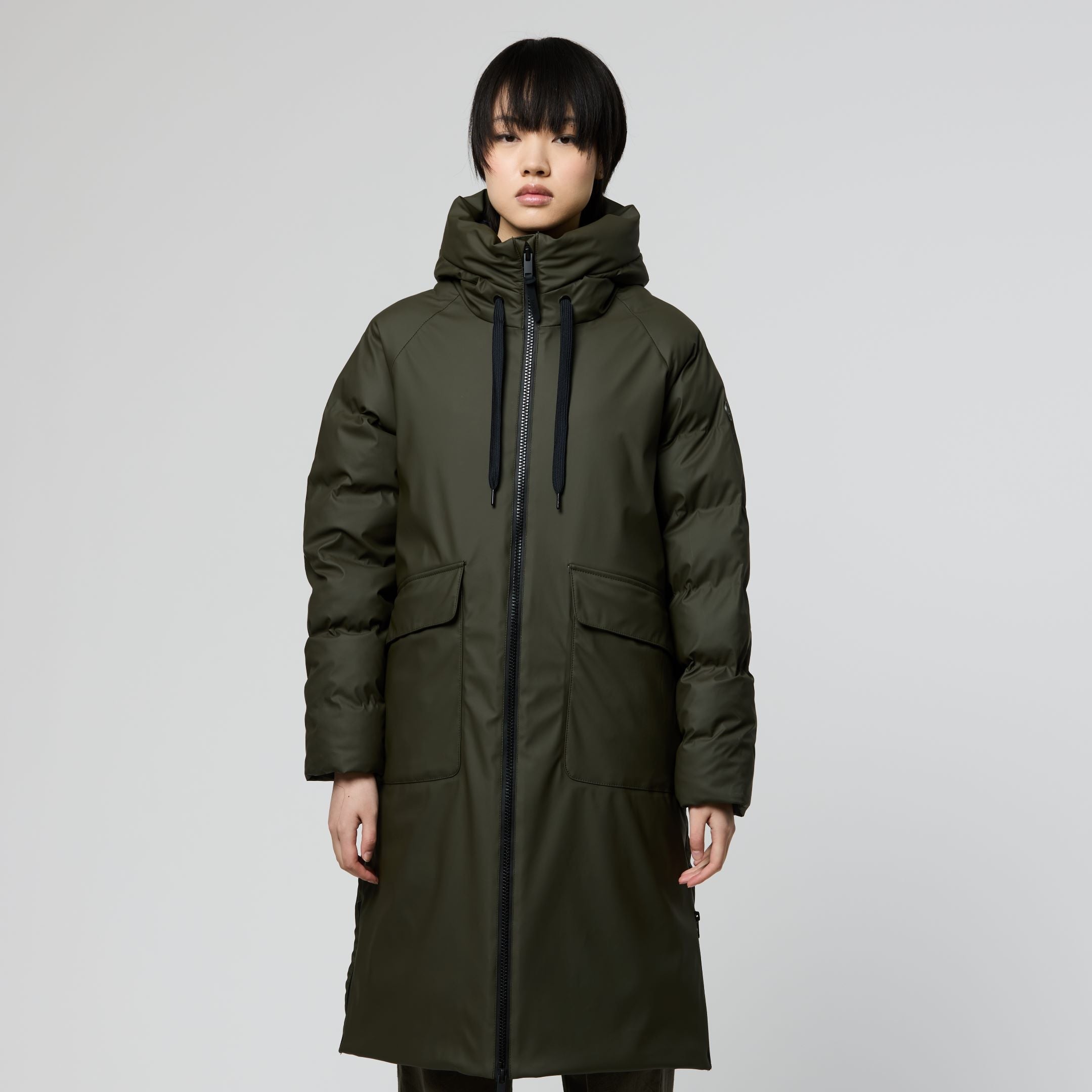 A model wearing a long, dark olive green puffer parka with a high collar, front zipper, and large front pockets, paired with matching dark trousers and brown shoes, set against a plain light gray studio background.