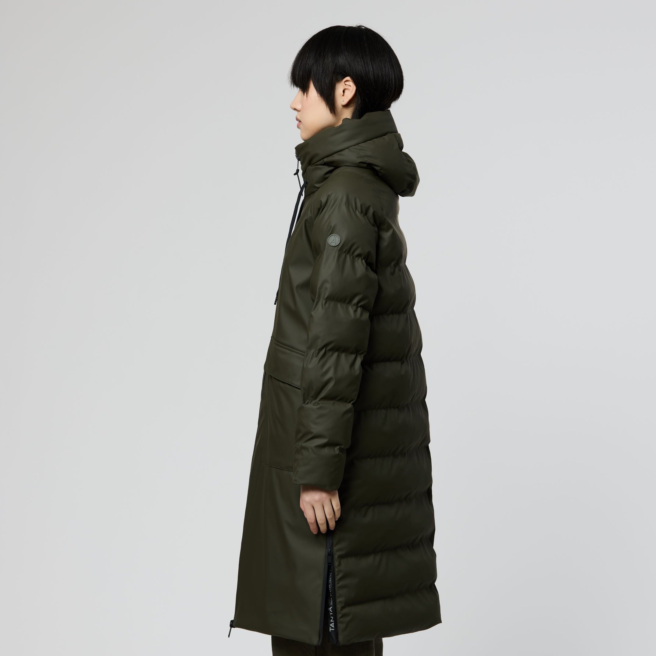 A model shown from the side profile wearing a long, dark olive green puffer coat, matching dark trousers, and brown suede boots against a light gray studio background.