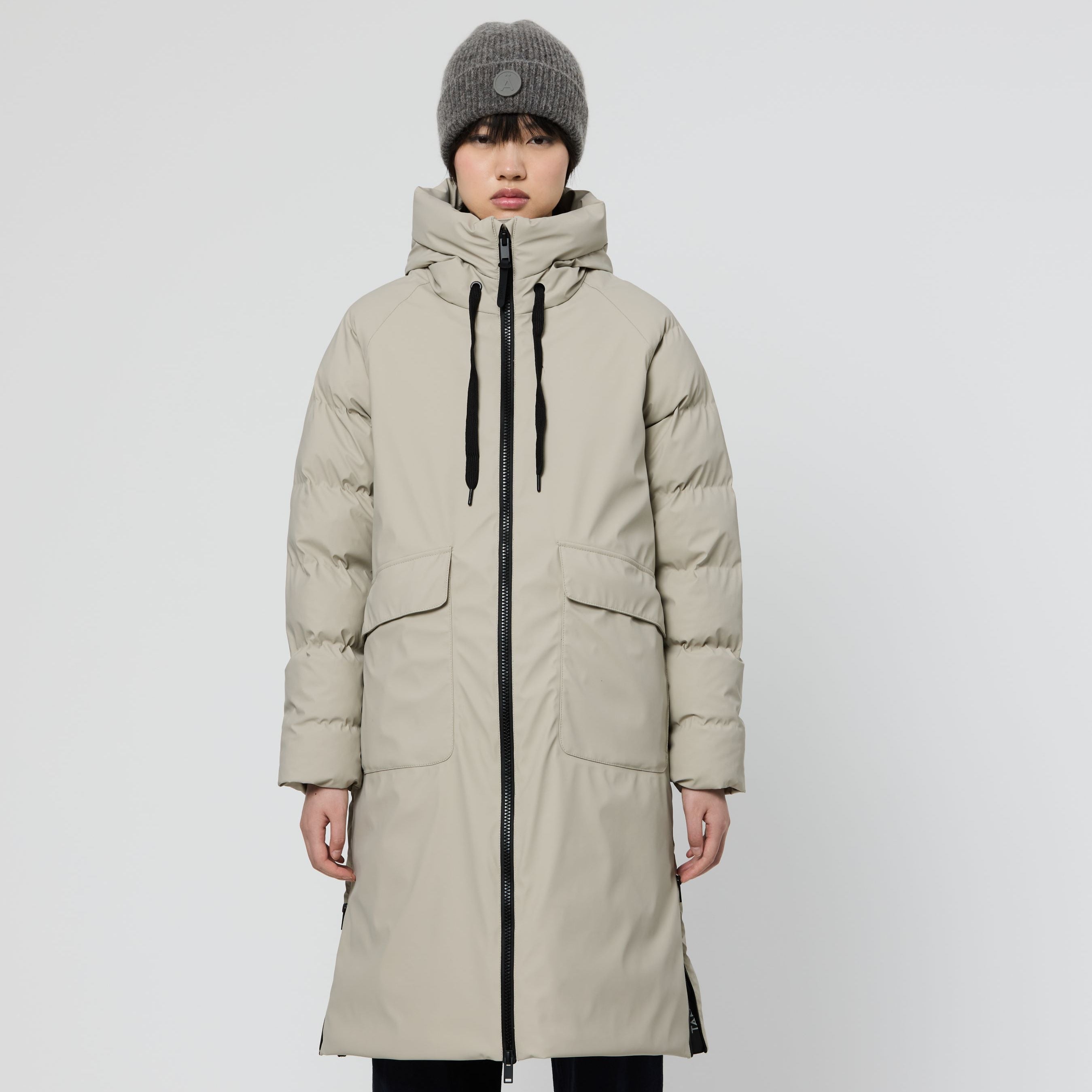 A model stands against a light gray studio background wearing a long, light beige puffer parka with black zippers and drawstrings, dark navy wide-leg pants, and a gray knit beanie.