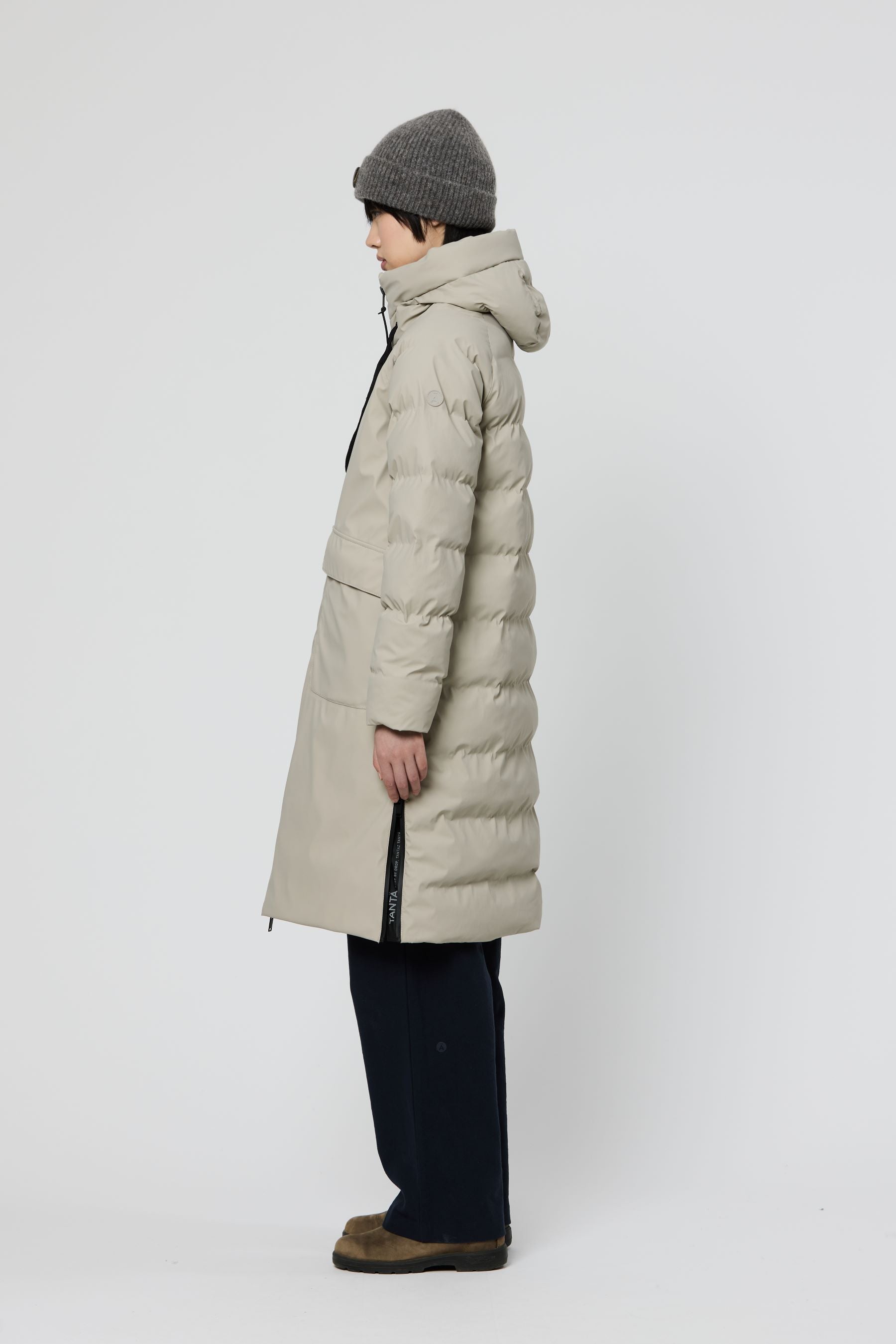 A person shown in side profile wearing a long puffer coat, knit beanie, dark pants, and brown shoes against a plain light gray studio background.