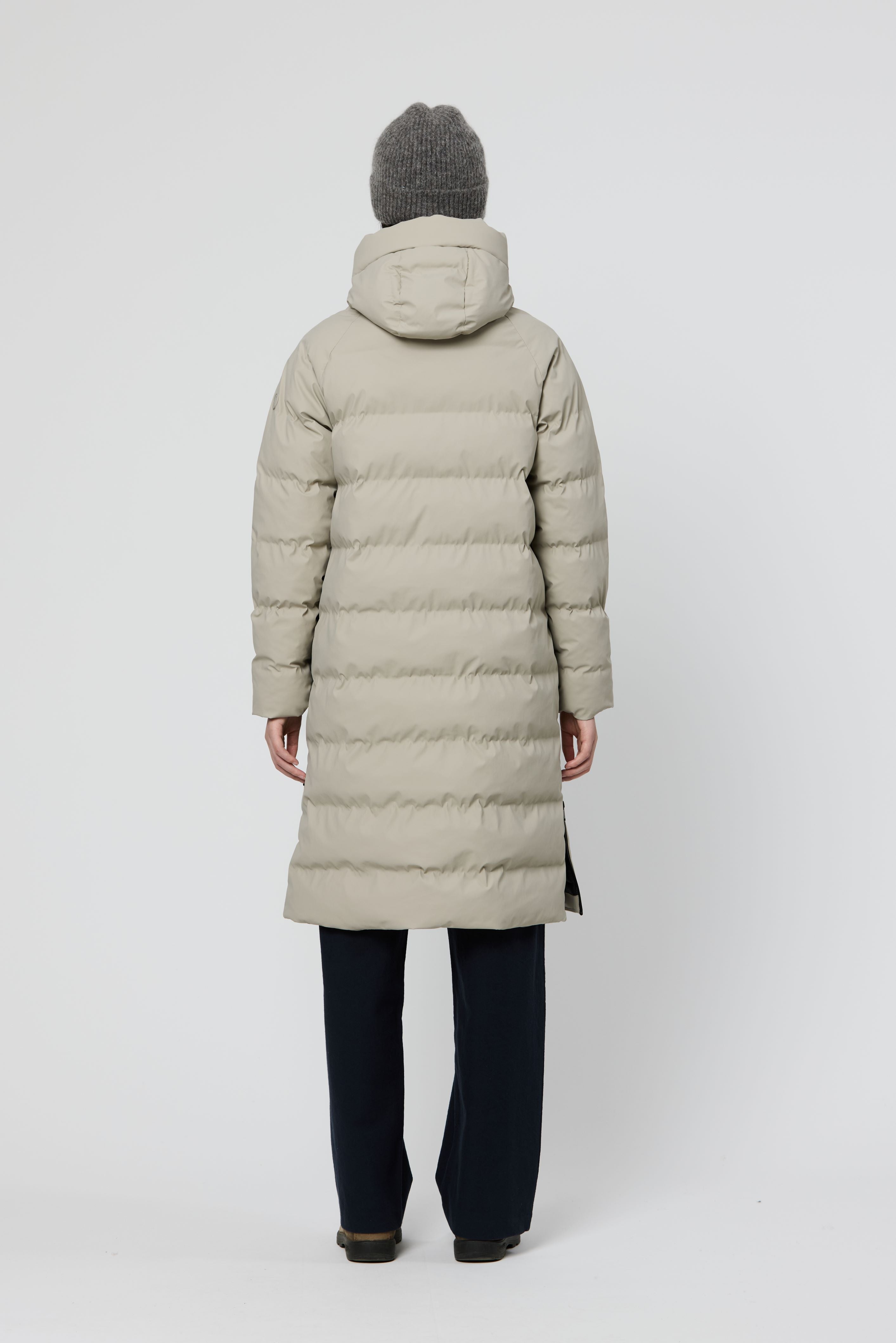 A person shown from the back wearing a long puffer coat in a light warm gray or greige color, with dark pants and a gray knit beanie, against a plain light gray studio background.