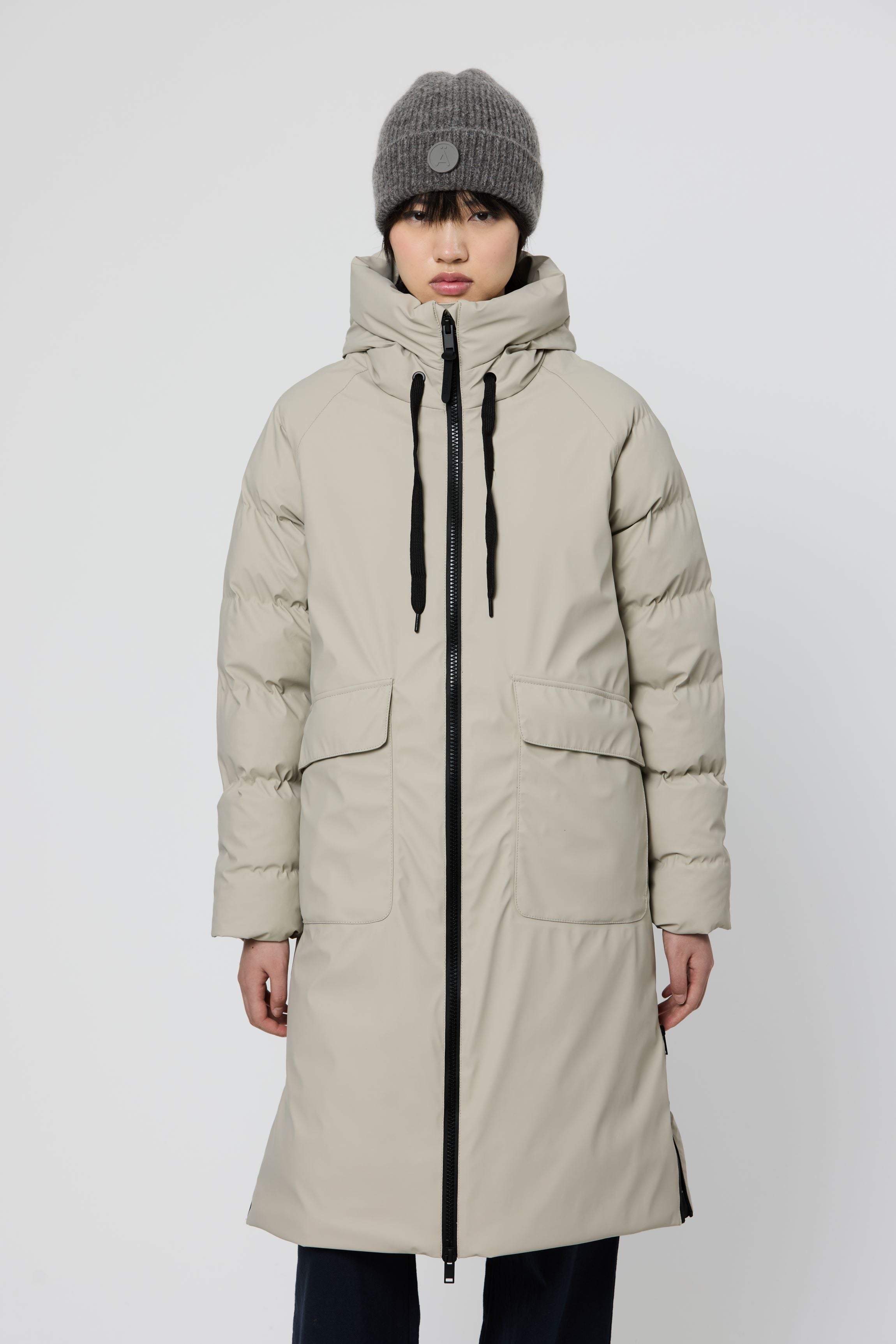 A model wearing a long, light beige hooded puffer coat with black zippers and drawstrings, paired with a gray knit beanie, against a plain light gray studio background.