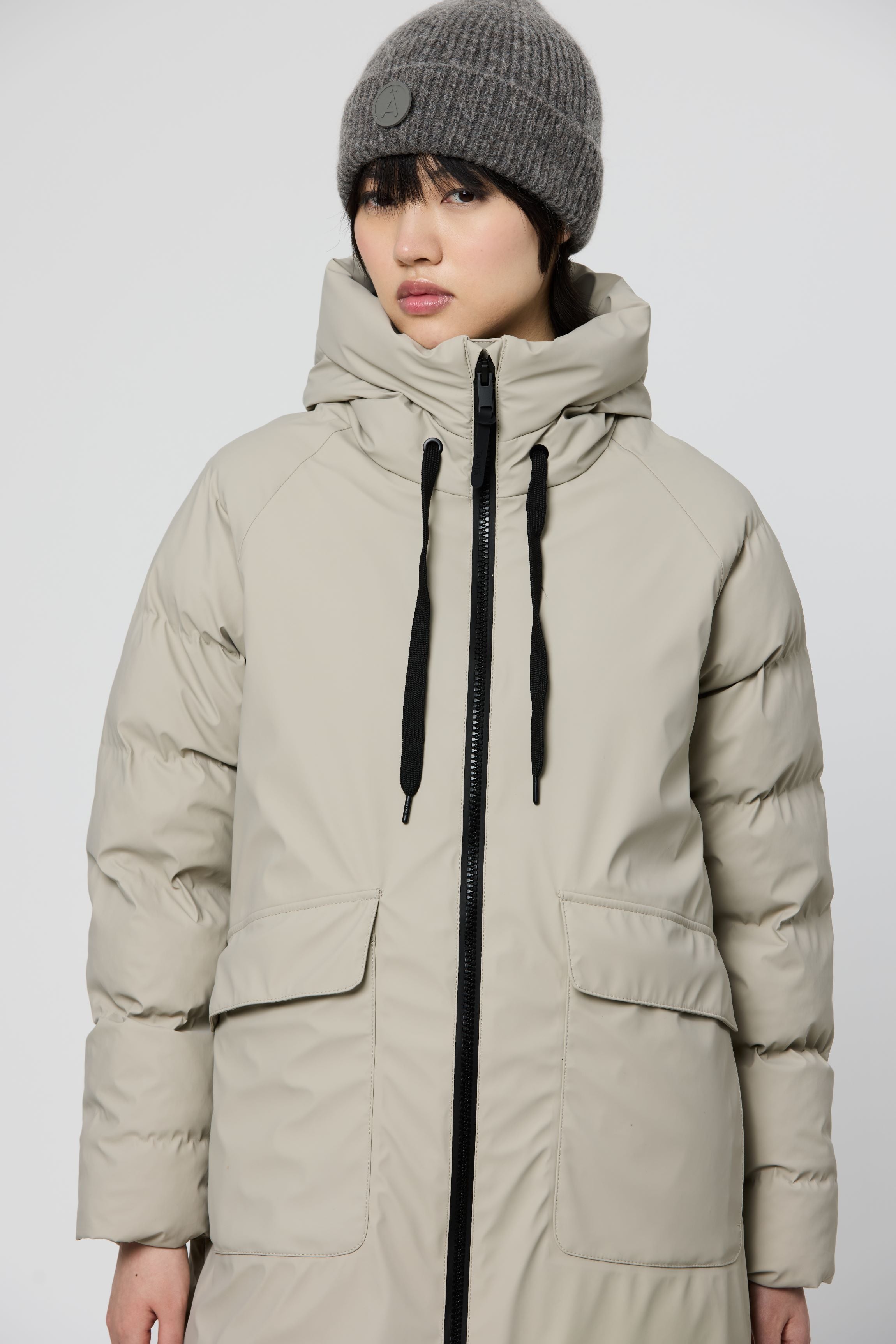 A model wearing a light beige hooded puffer coat with a black front zipper and black drawstrings, paired with a gray knit beanie, against a plain light gray studio background.