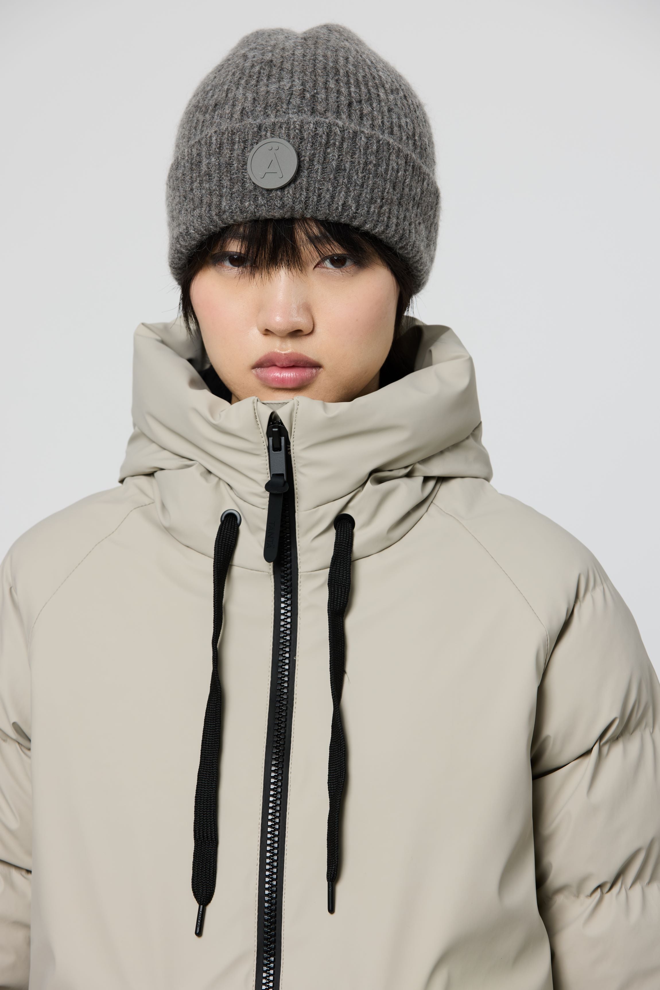 A model wearing a light beige hooded puffer jacket with black drawstrings and a gray ribbed knit beanie hat featuring a circular button detail, shot against a light gray studio background.