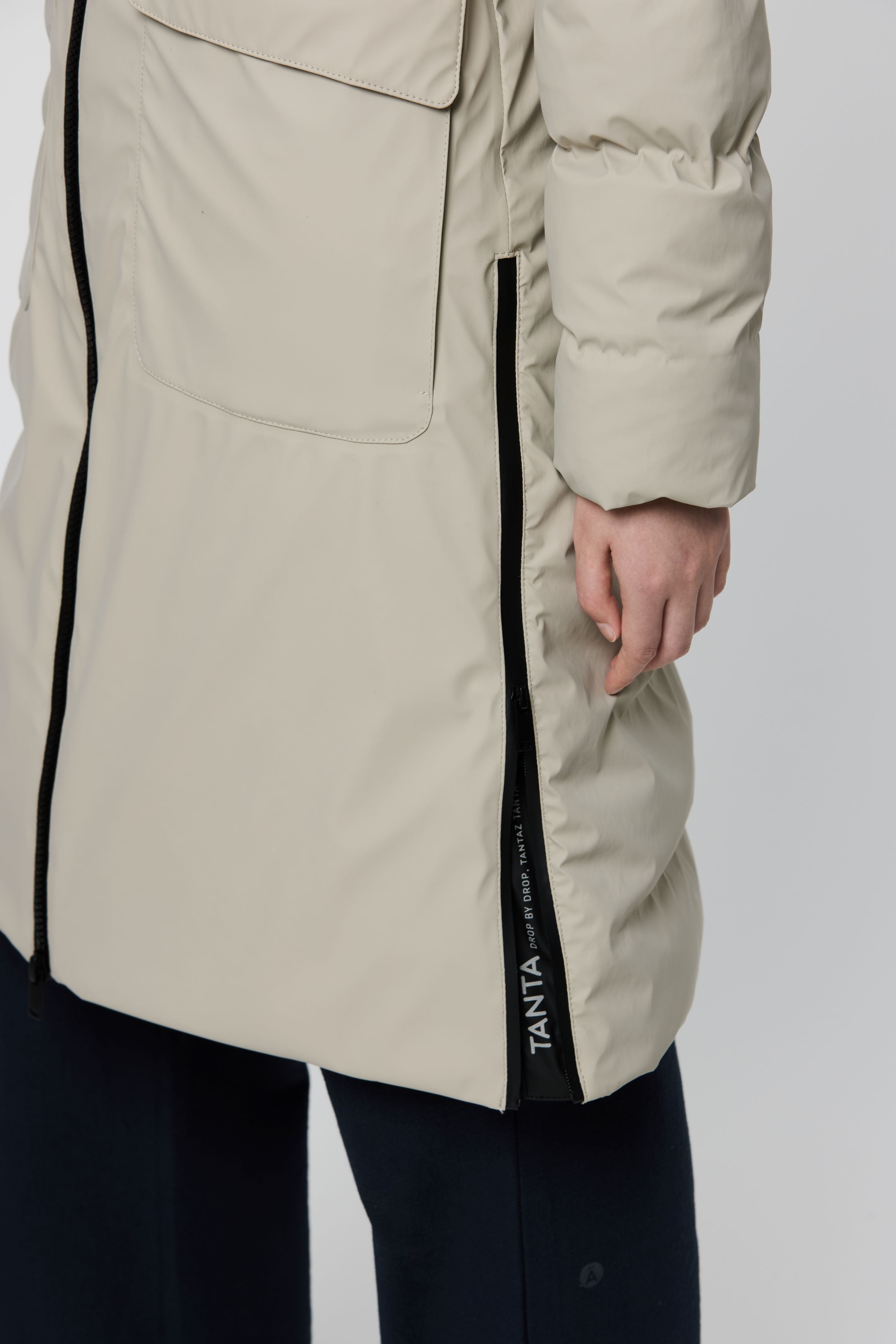 A close-up of a person wearing a light beige puffer coat with black side trim and a vertical TANTA logo, paired with dark pants, against a light gray studio background.