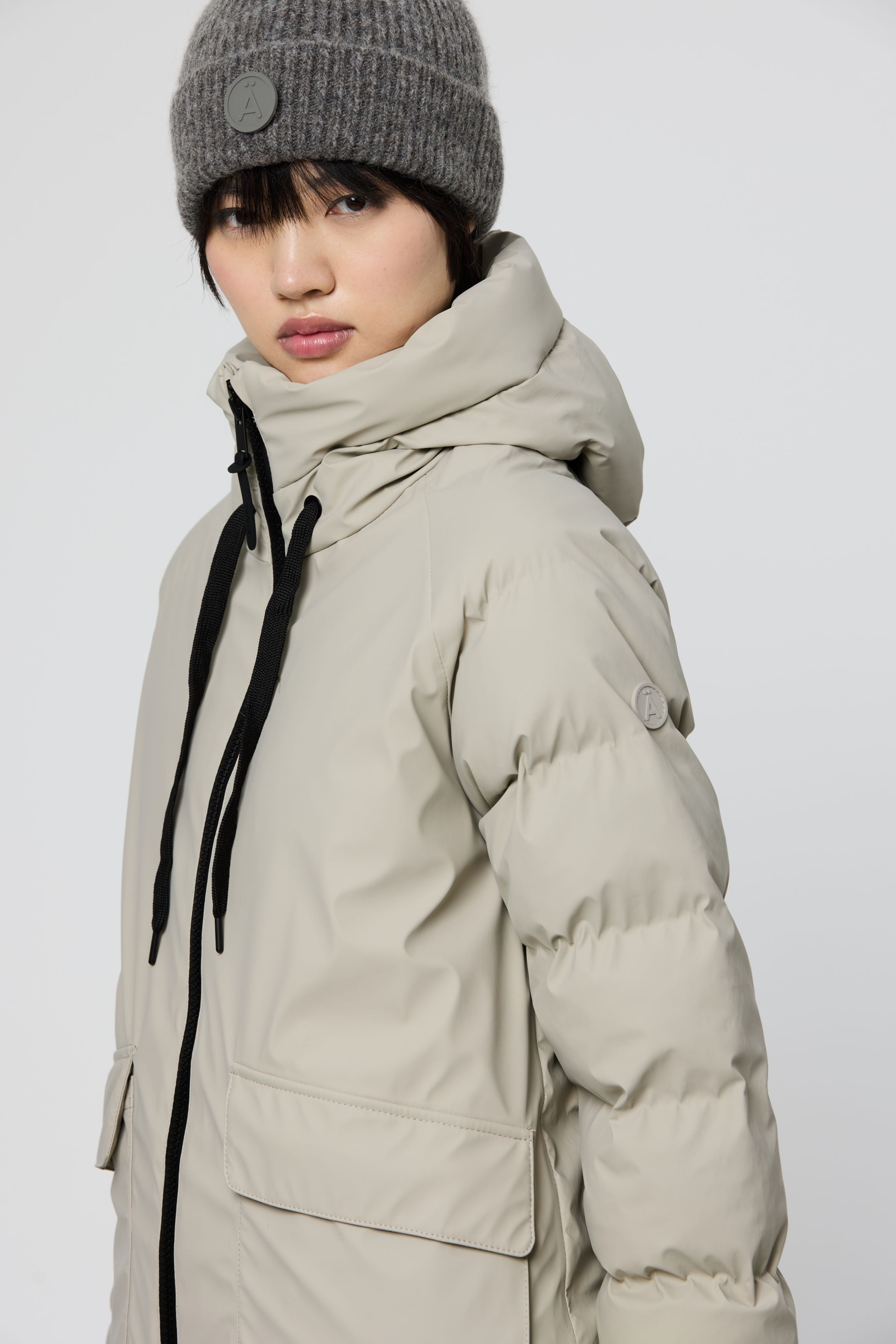 A model wearing a light beige hooded puffer jacket with black drawstrings and a gray knit beanie, shown in a studio setting against a light gray background.