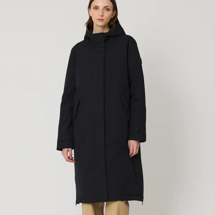 A model stands against a light gray studio background wearing a long black hooded coat, beige wide-leg trousers, and dark brown shoes.