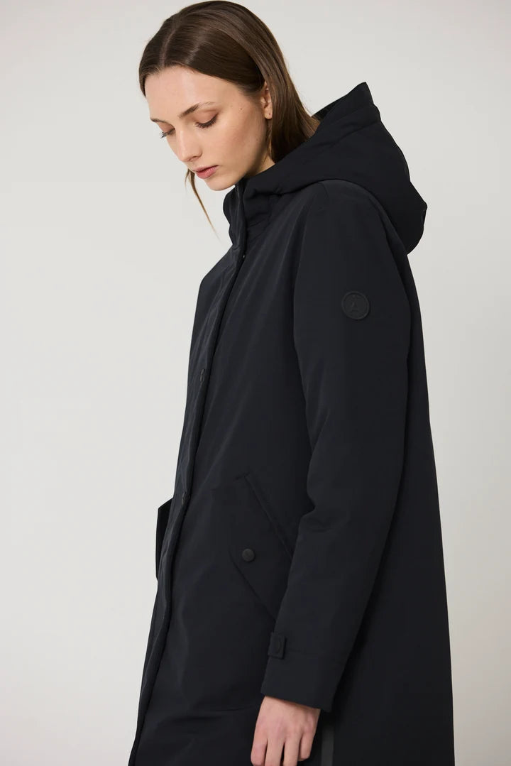A woman wearing a long black hooded coat, shown in profile against a plain light gray studio background.