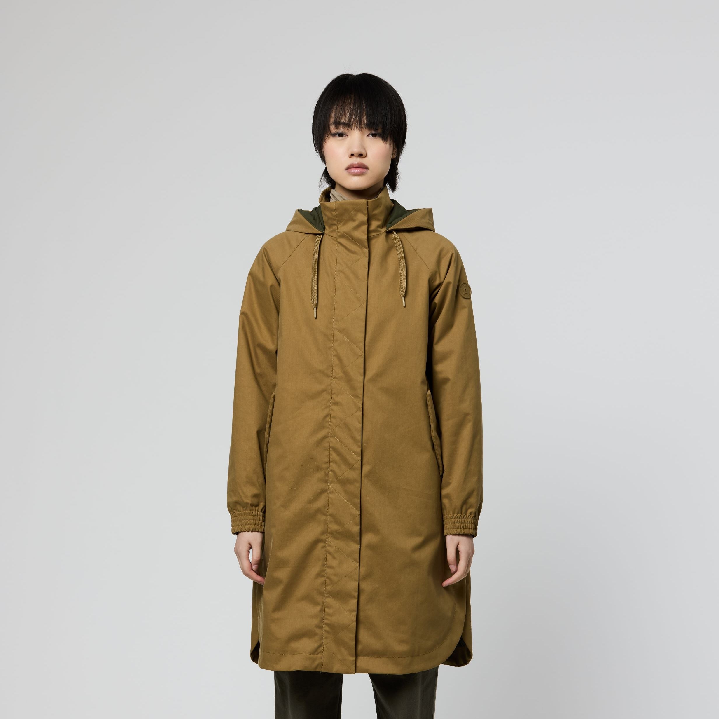 A model stands against a light gray studio background wearing a mid-length hooded parka in a warm camel-brown color, paired with dark olive pants and brown shoes.