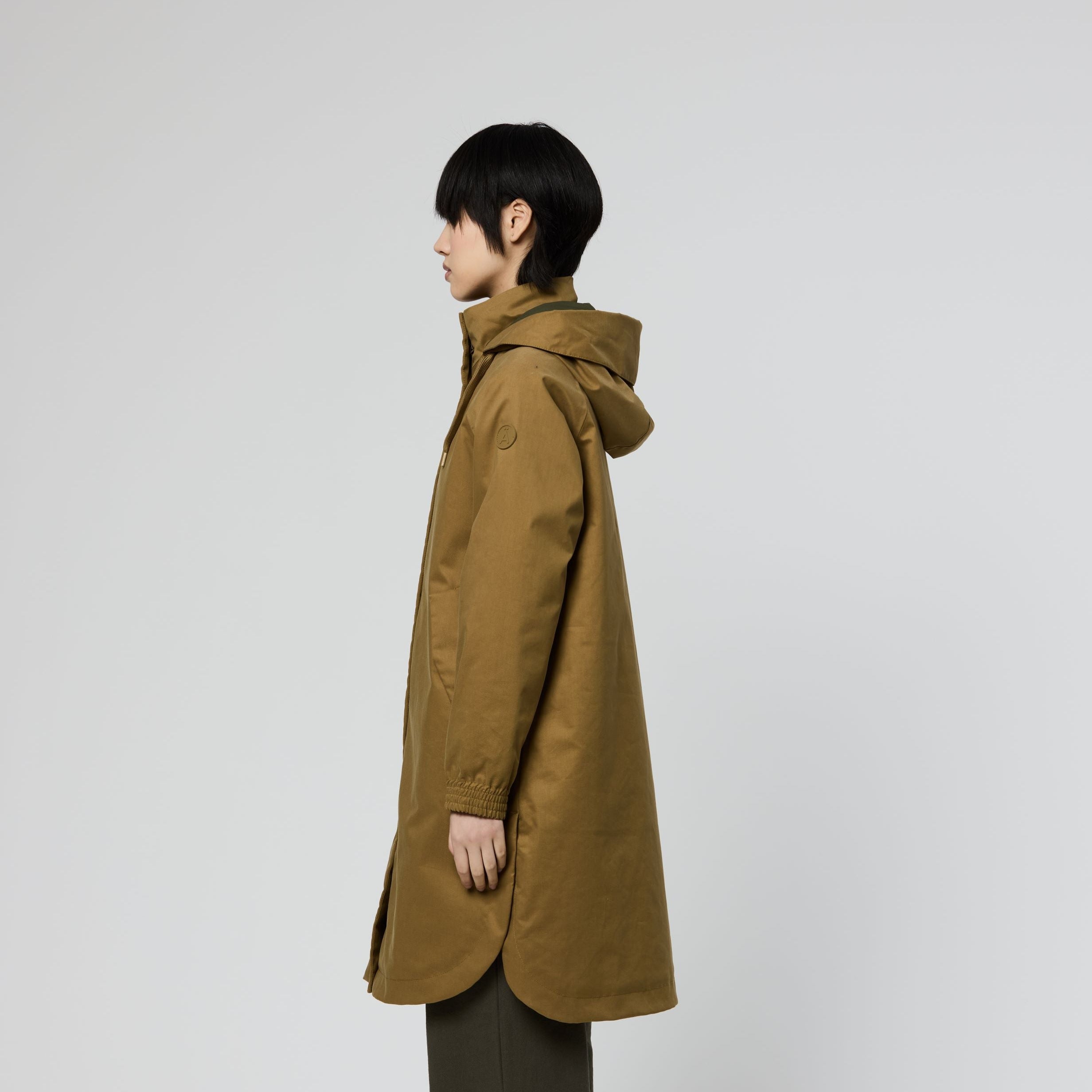 A model shown in profile wearing a mid-length olive-brown hooded coat and dark olive pants against a plain light gray studio background.