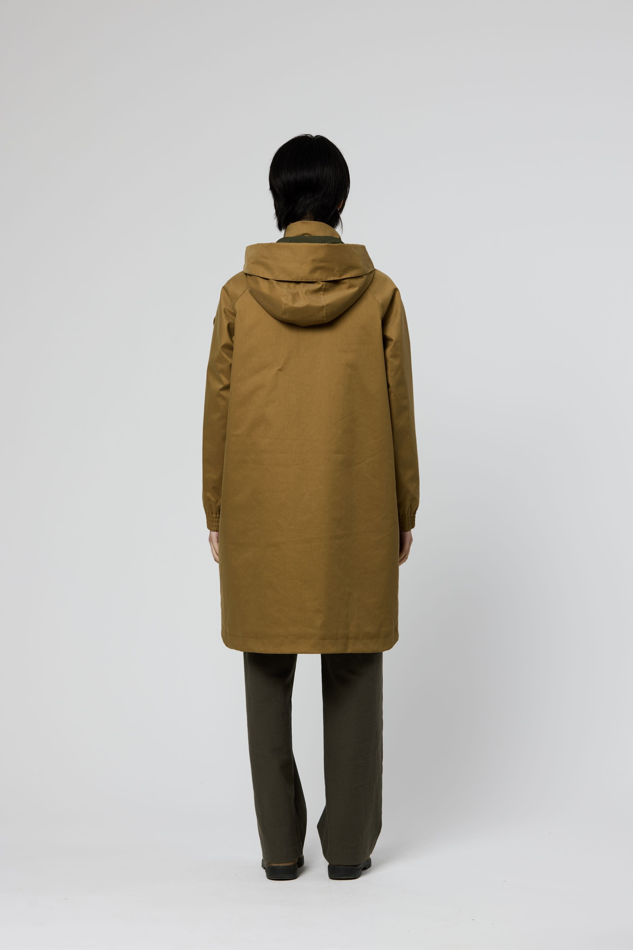 A person shown from the back wearing a mid-length hooded coat in a warm camel-brown color, paired with dark olive pants, against a plain light gray studio background.