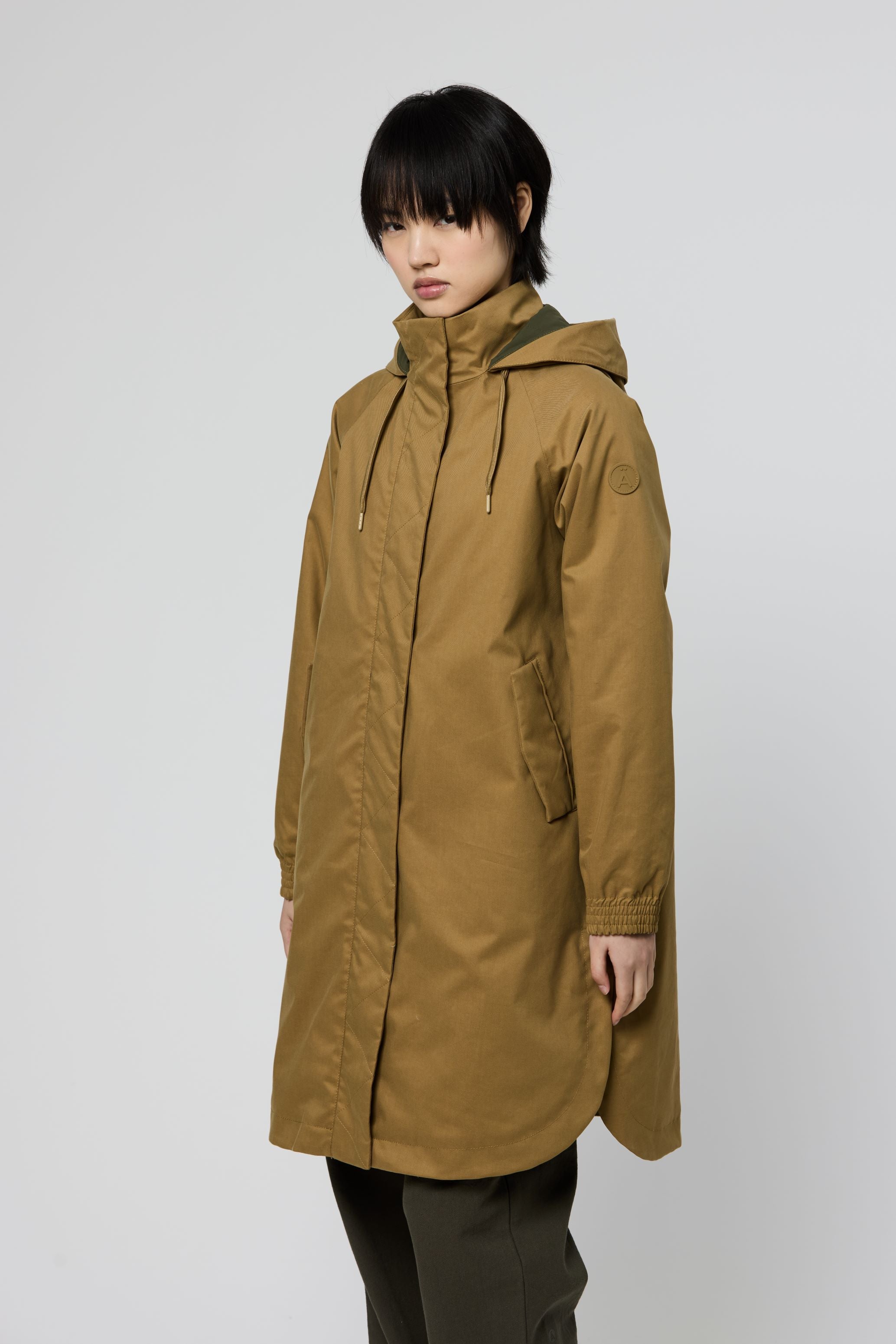 A model wearing a mid-length hooded parka coat in a warm camel brown color, paired with dark pants, photographed against a neutral light gray studio background.