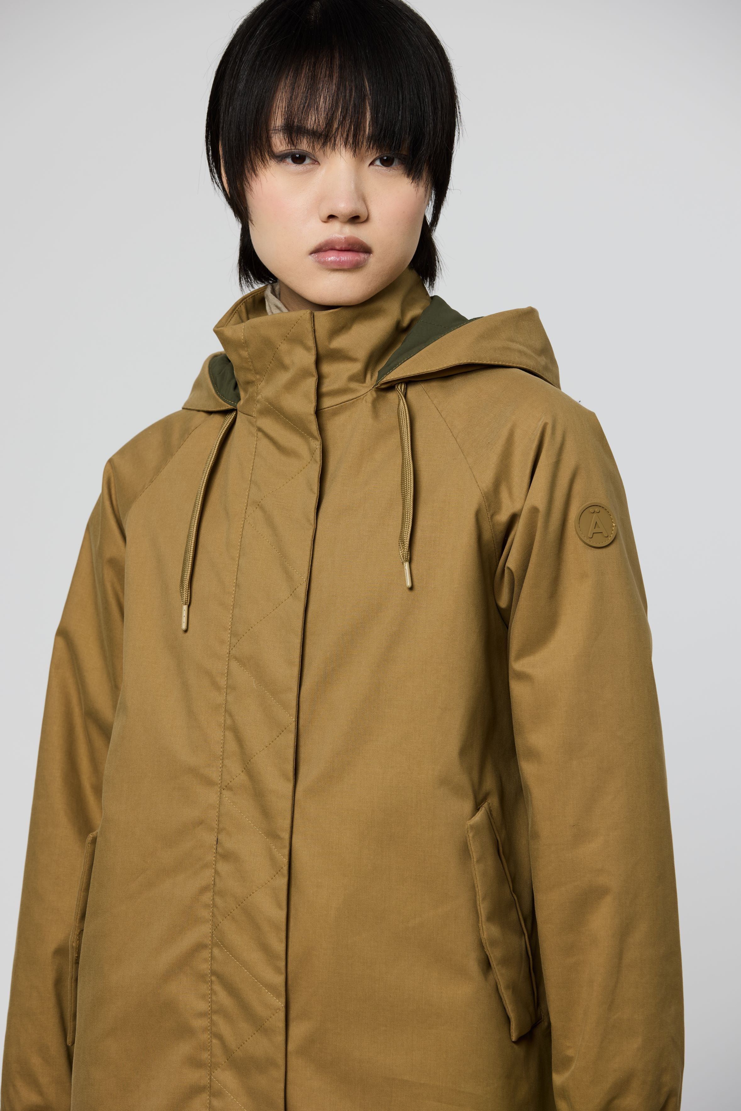 A model wearing a hooded jacket in a warm camel brown color, featuring a high collar, front zipper, and drawstrings, set against a plain light gray background.