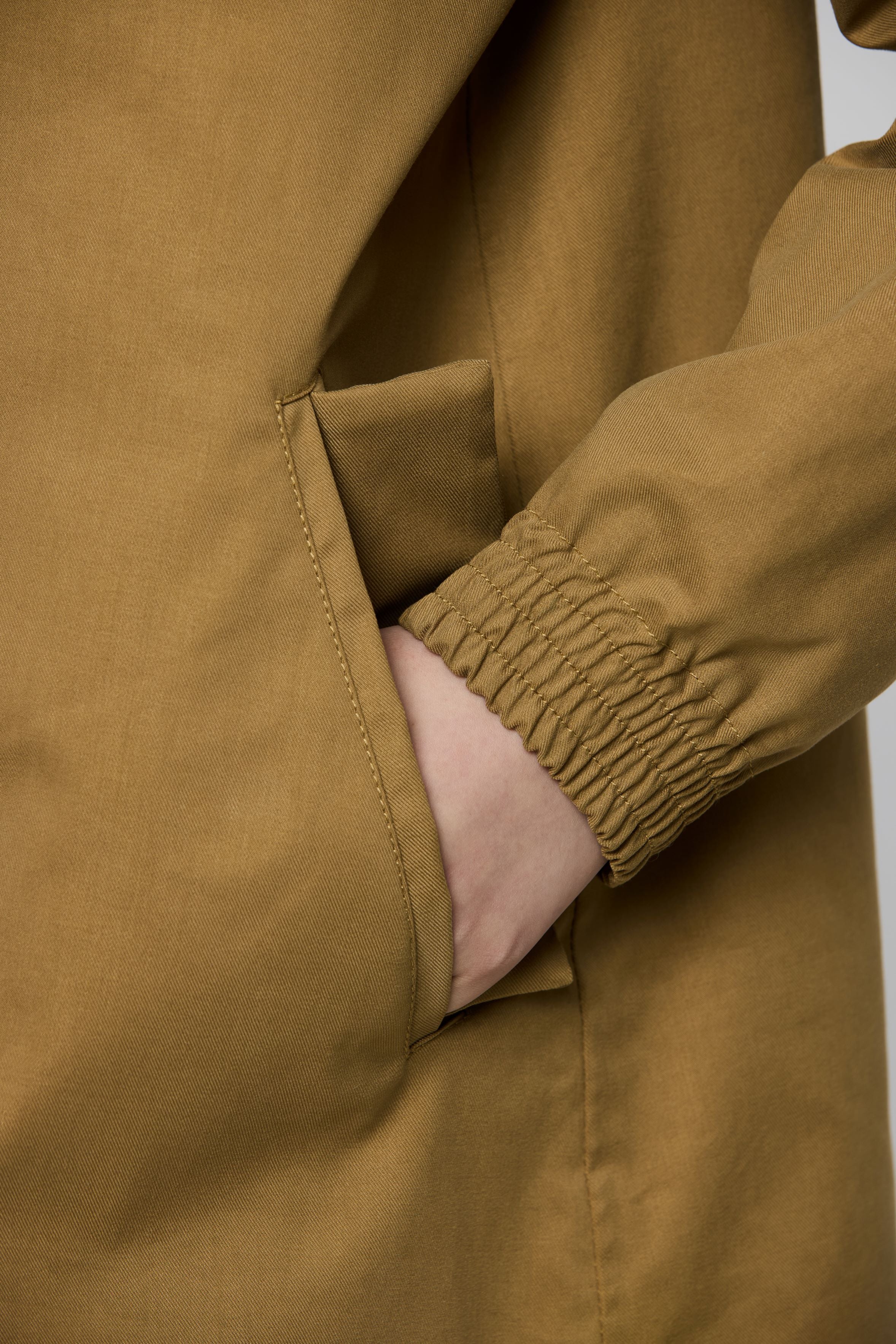 Close-up of a person wearing a camel-colored jacket, showing a side pocket with a hand tucked inside and a gathered elasticated cuff on the sleeve.