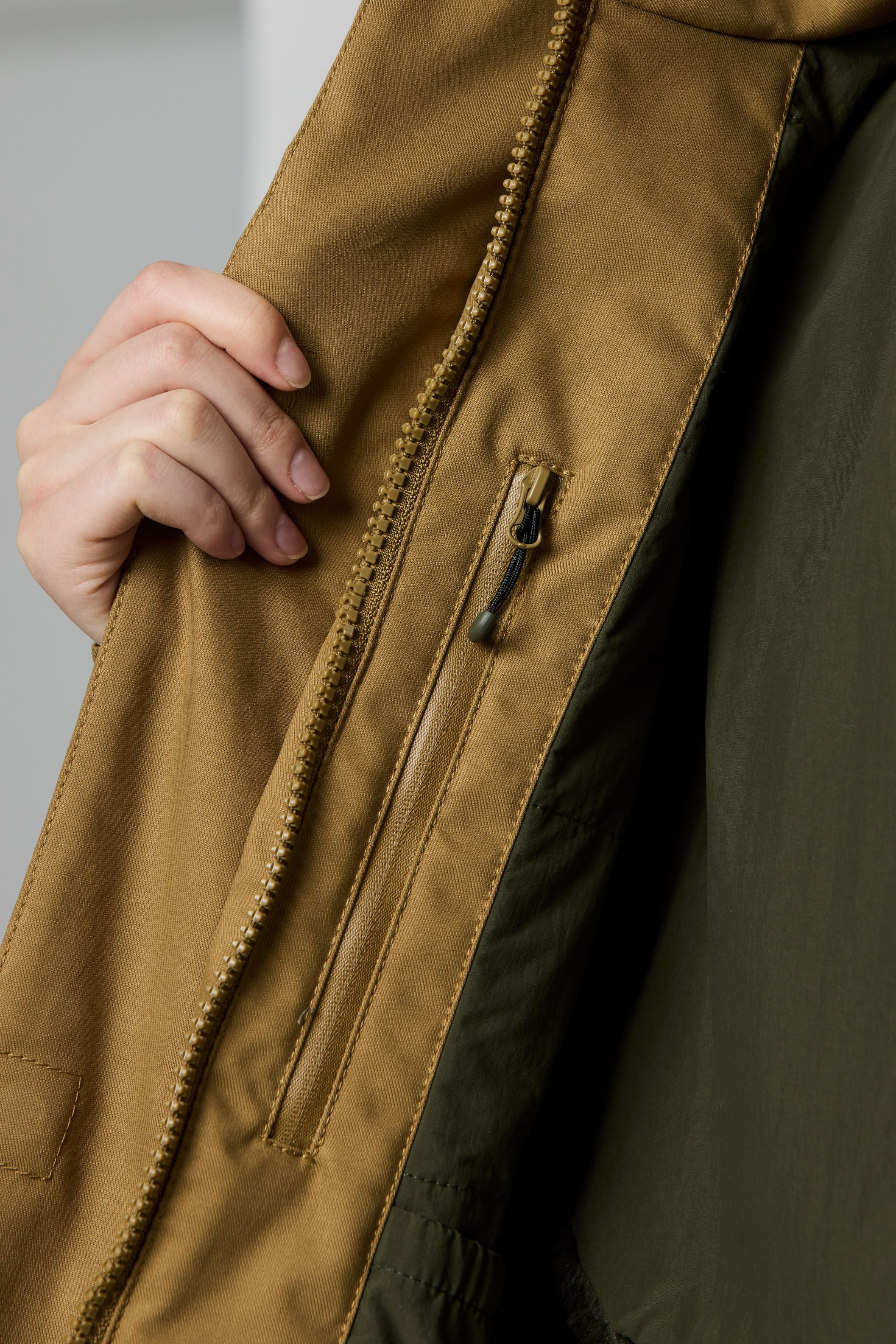 Close-up of a person wearing a jacket, showing a tan outer panel with a vertical zip pocket and a dark olive green inner panel, with a hand holding the tan fabric open.