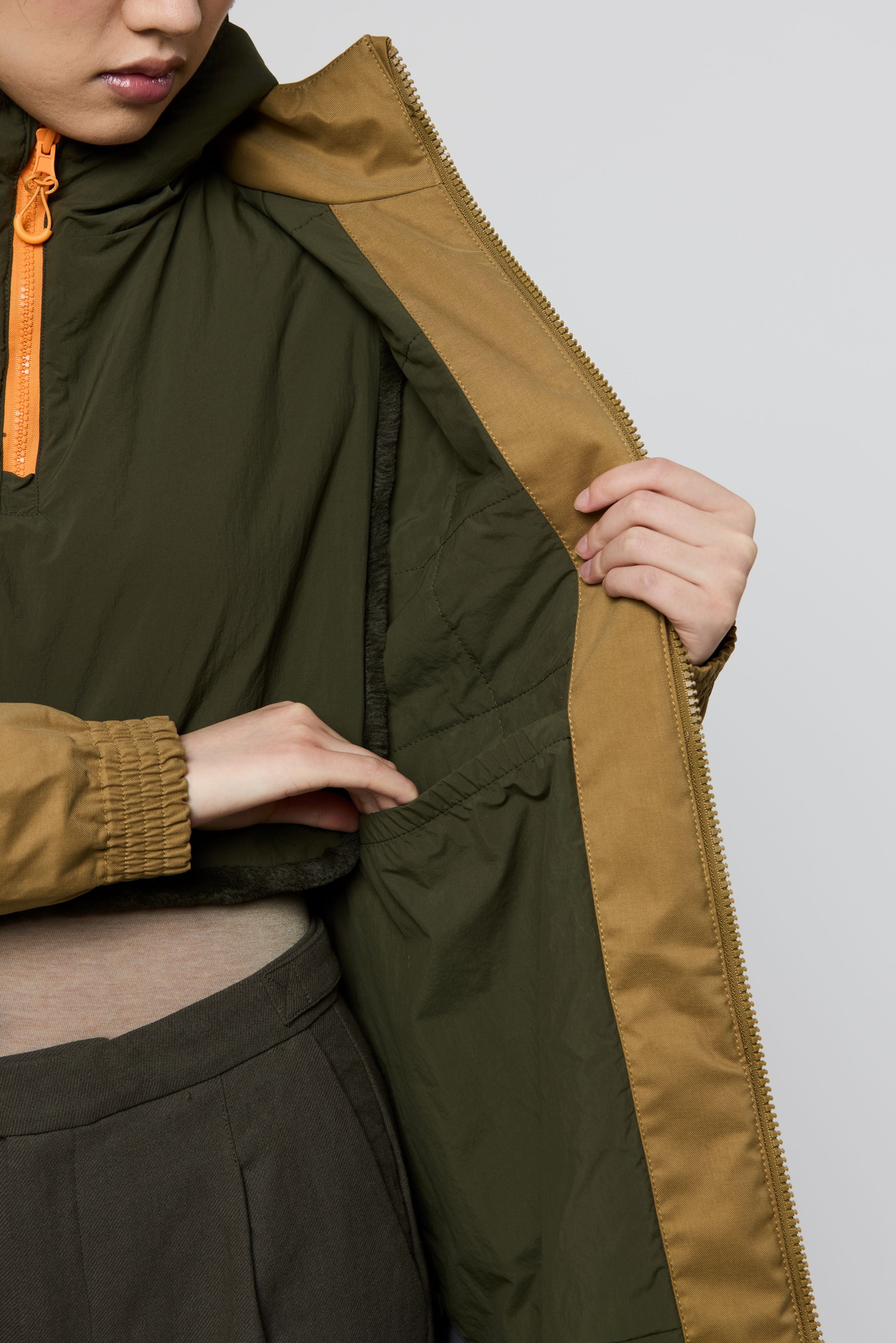 Close-up of a person wearing a hooded jacket with an olive green exterior and a camel-brown interior lining, holding the jacket open to show the lining. The person is also wearing dark trousers and a beige top.