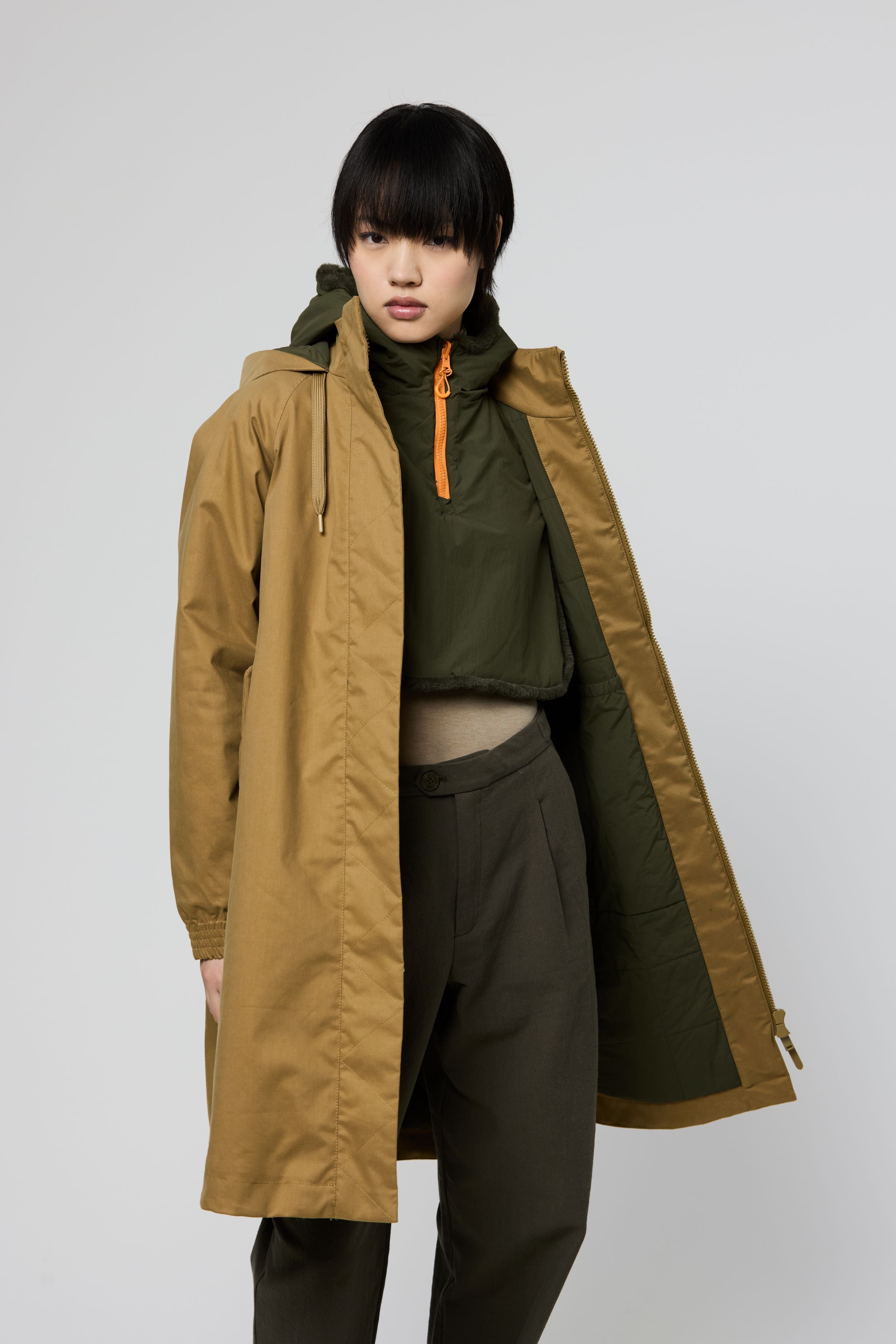 A model wearing a long tan hooded jacket with a dark olive green inner lining and a bright orange zipper, styled over an olive green crop top and dark pants against a light gray studio background.