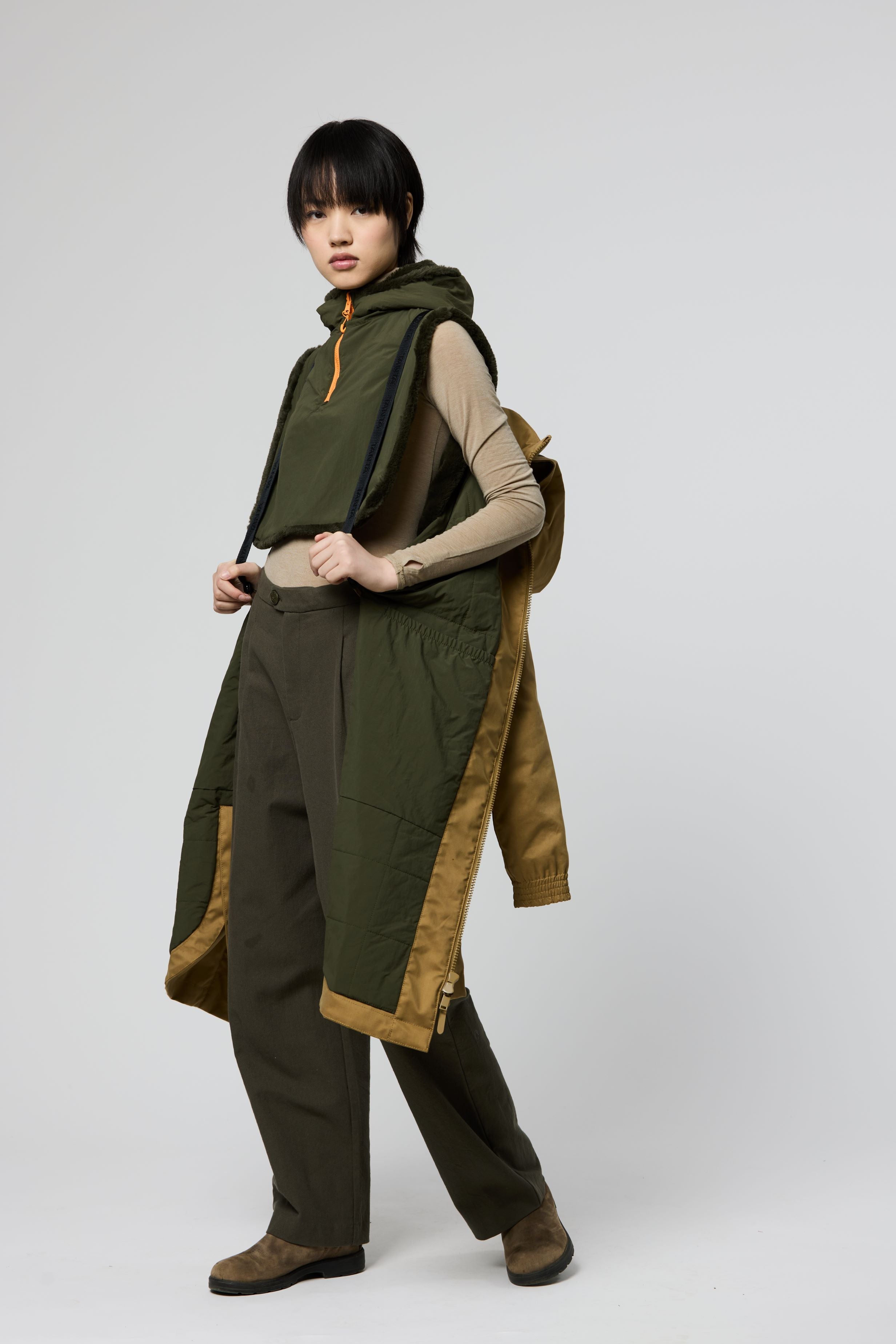 A model stands against a light gray studio background wearing a long olive green hooded outerwear piece with tan contrast panels and dark pants.