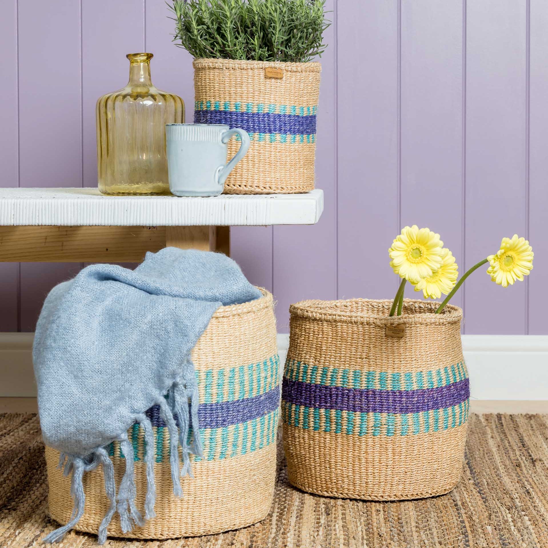 Three natural-fiber baskets with teal and purple stripes.  Two larger baskets are on the floor; one holds a light blue throw.