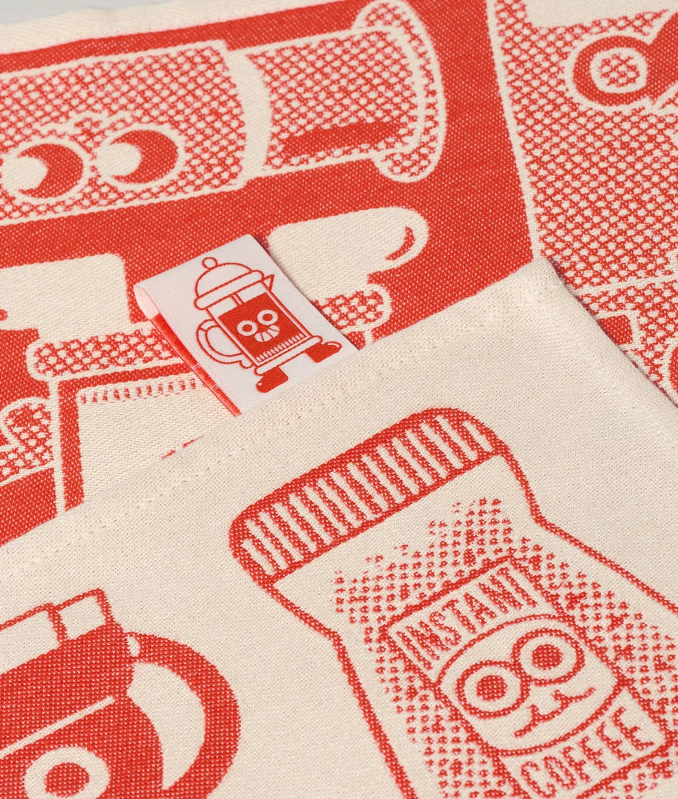 A close-up of a cream-colored fabric item, likely a pocket or pouch, featuring a red printed pattern of coffee-themed illustrations such as mugs, a coffee bag labeled 'INSTANT COFFEE,' and a small sewn fabric tag with a coffee cup graphic.
