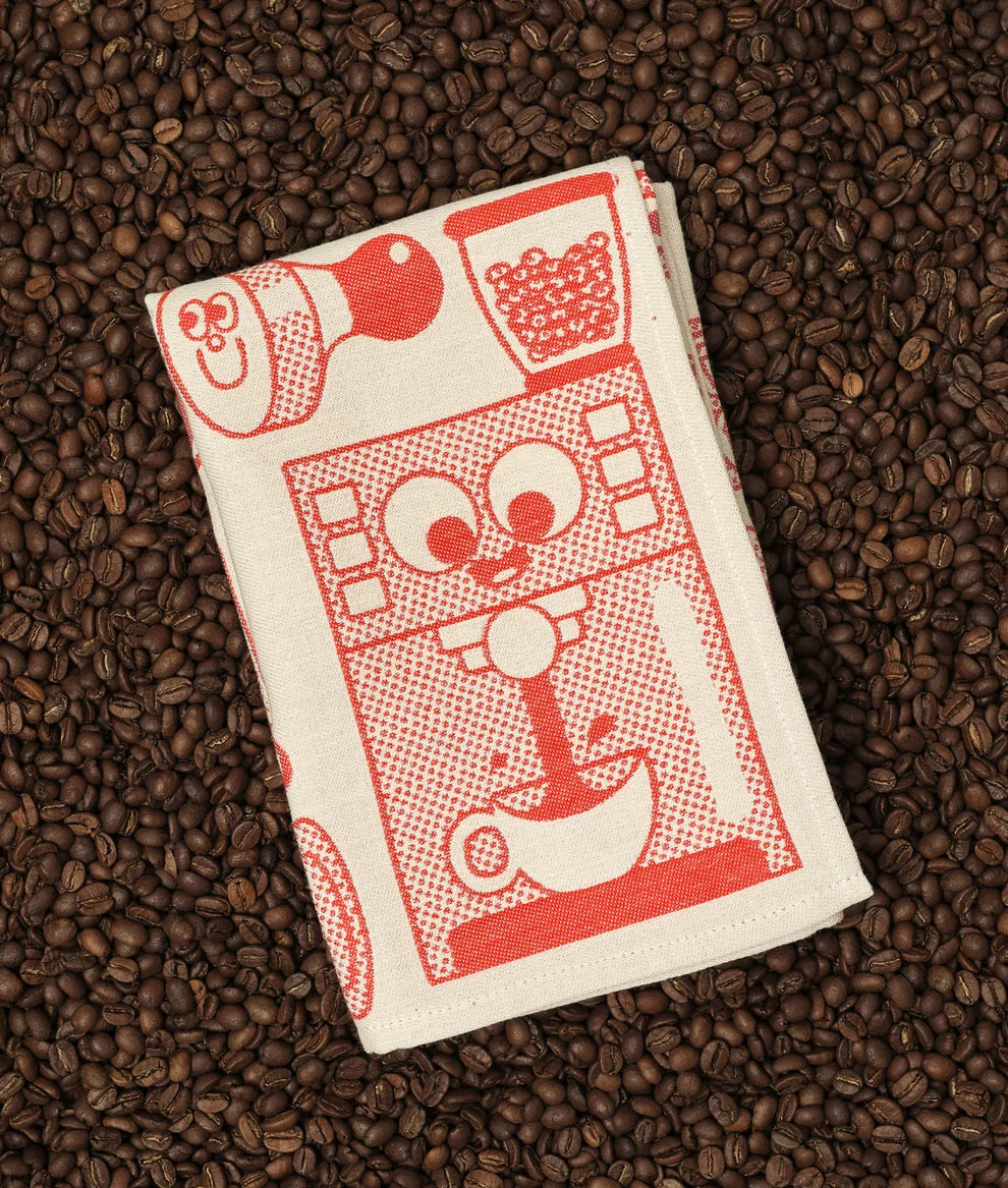 A folded white kitchen towel with a red graphic print of a coffee machine and coffee cups, placed on a bed of dark roasted coffee beans.