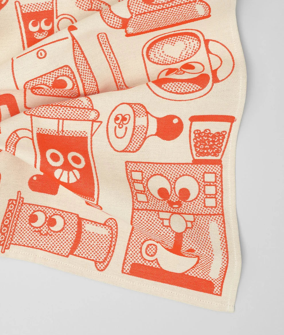 A close-up of a cream-colored fabric item, likely a tea towel or napkin, featuring a retro-style orange graphic print of coffee and tea cups, mugs, and appliances with cartoon faces.