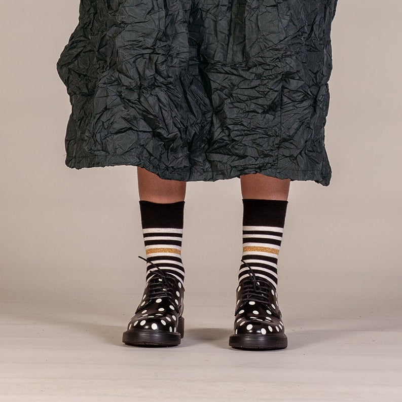 A person wearing a dark crinkled midi skirt, black and white striped socks with a thin gold stripe, and black lace-up boots with white polka dots, standing against a neutral beige studio background.