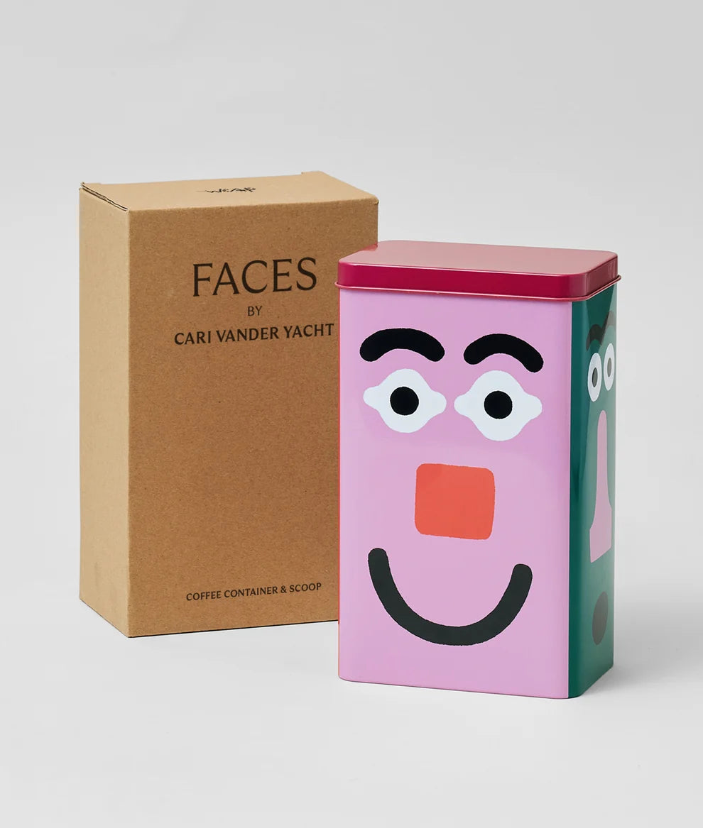A pink metal tin with a cartoon face design stands in front of its brown cardboard box. The box is printed with the text 'FACES BY CARI VANDER YACHT' and 'COFFEE CONTAINER & SCOOP'.