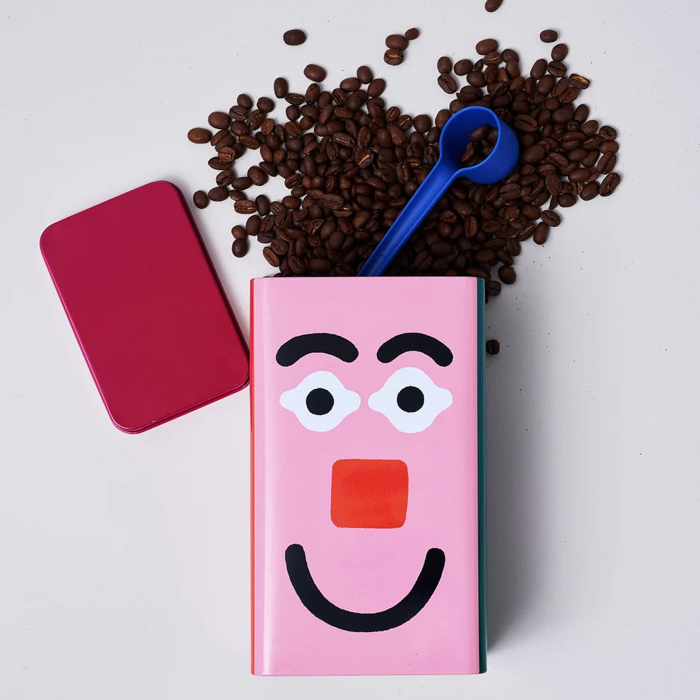 Pink container with cartoon face design, surrounded by coffee beans and a blue scoop on a light gray background