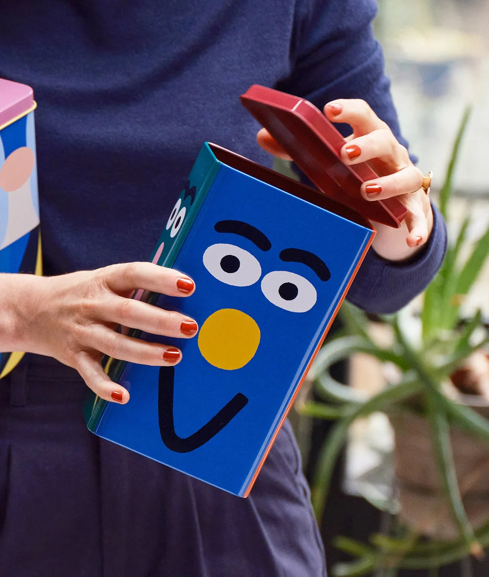 A person holding a colorful blue tin canister designed to look like the character Grover from Sesame Street, with the lid propped open. The background is a softly blurred indoor setting with a plant.