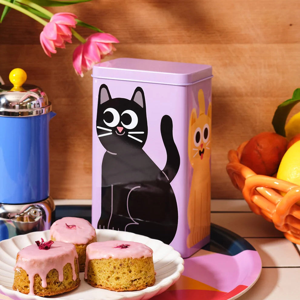 Purple tin box with cartoon cat design on a wooden surface with pastries and flowers.