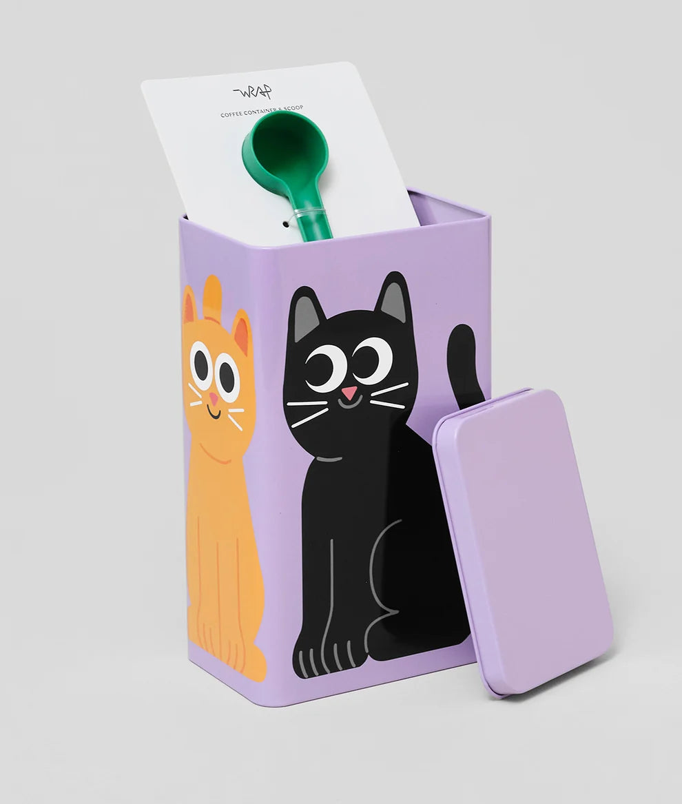 A lavender metal tin with a matching lid, decorated with cartoon cat illustrations in orange and black. A green spoon and a white card labeled 'WAP' are visible inside the tin, set against a plain light gray background.