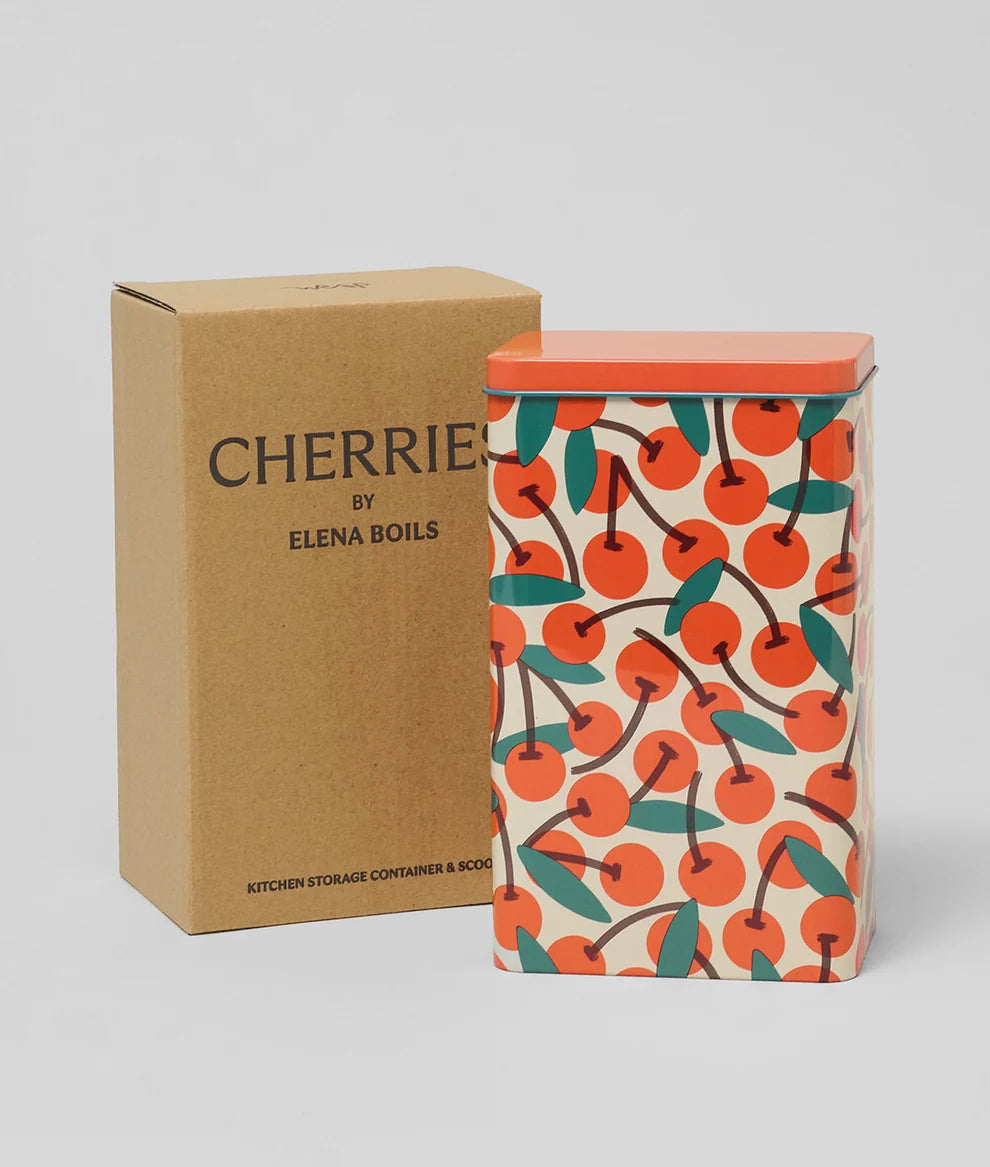 A rectangular metal canister with a bright orange lid and a cream background covered in an orange cherry print, standing next to its brown kraft paper box labeled 'CHERRIES BY ELENA BOILS' on a light gray background.