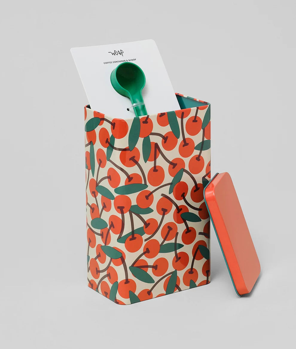 A patterned paper shopping bag with an orange and green cherry print, shown with a green spoon inside and a matching orange lid leaning against it on a light gray background.
