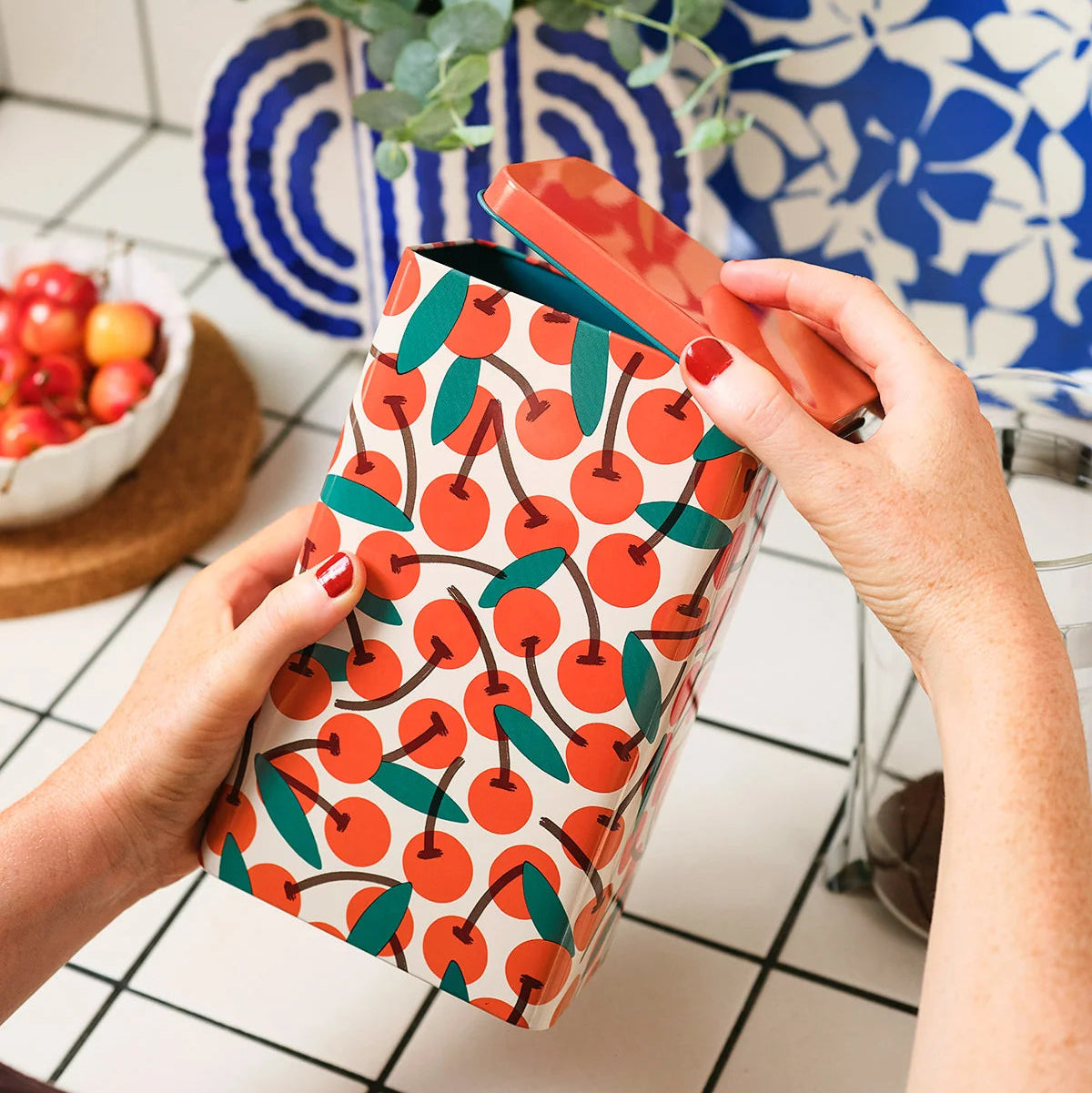 A person's hands with red nail polish hold a white paper gift bag decorated with a vibrant orange and green cherry pattern, set against a tiled kitchen background.