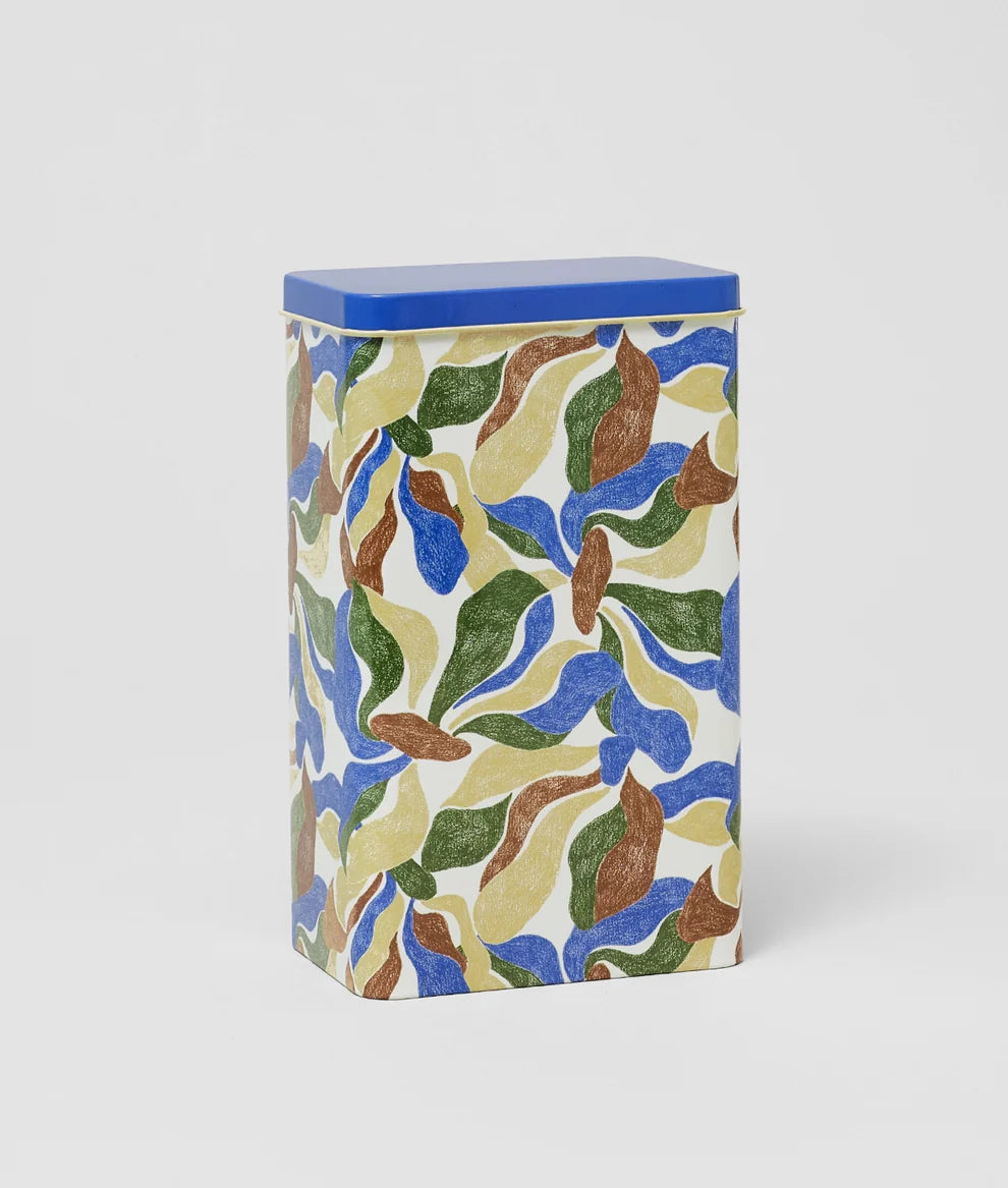 A rectangular metal tin with a blue lid and a colorful abstract leaf pattern in blue, green, brown, and cream on a light background, shown on a neutral light gray studio background.