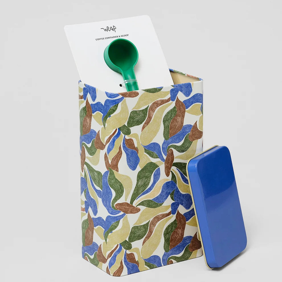 A patterned storage box with a blue lid leaning against it, shown on a light gray studio background. The box has an abstract leaf-like print in blue, green, brown, and cream, and a green scoop or spoon-like tool is partially visible inside, with a white card protruding from the top.