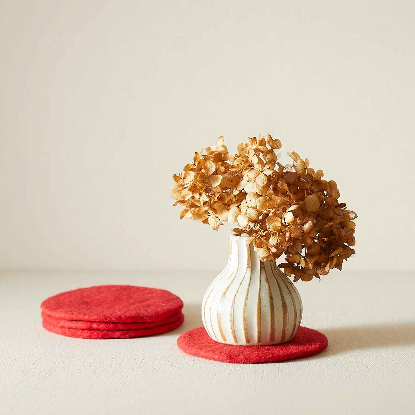 Dried hydrangea in a vertically ribbed, off-white vase with gold accents, sitting on a red felt coaster.