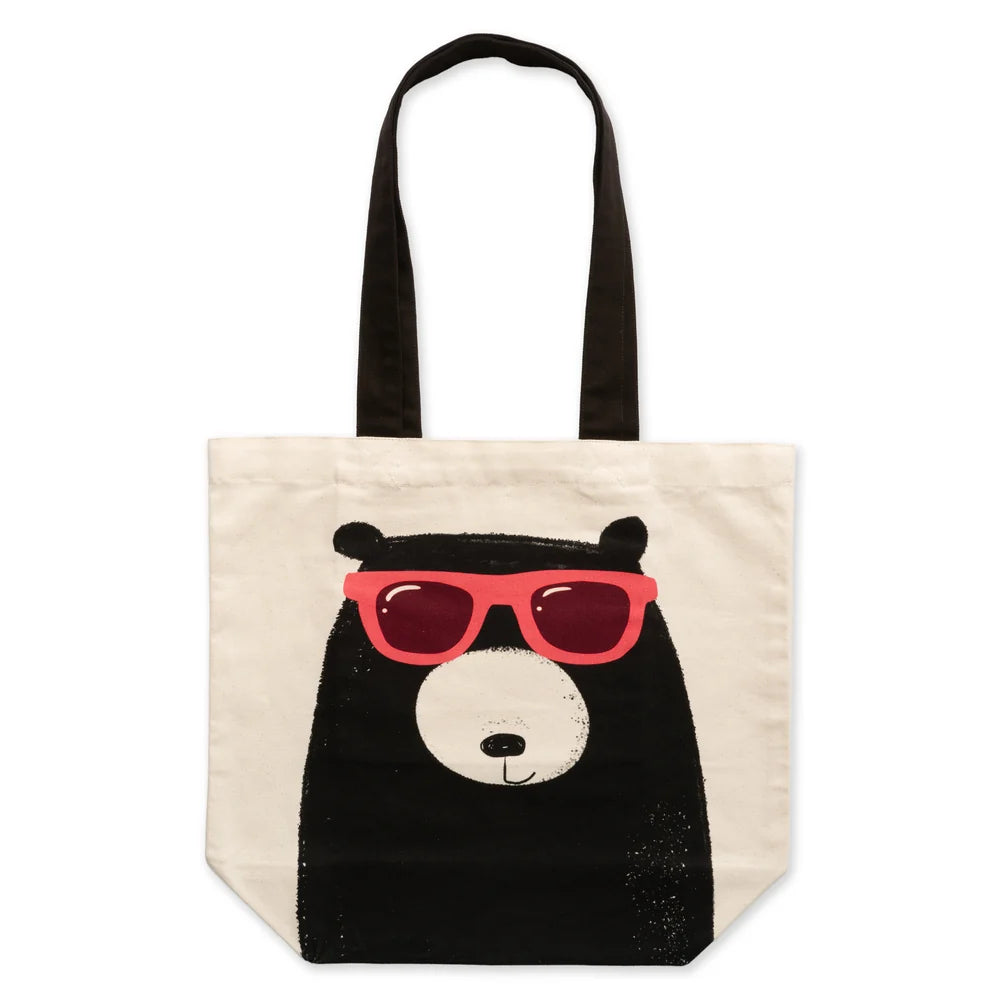 A cream canvas tote bag with black shoulder straps, featuring a graphic print of a black bear face wearing bright pink sunglasses on the front, set against a plain white background.