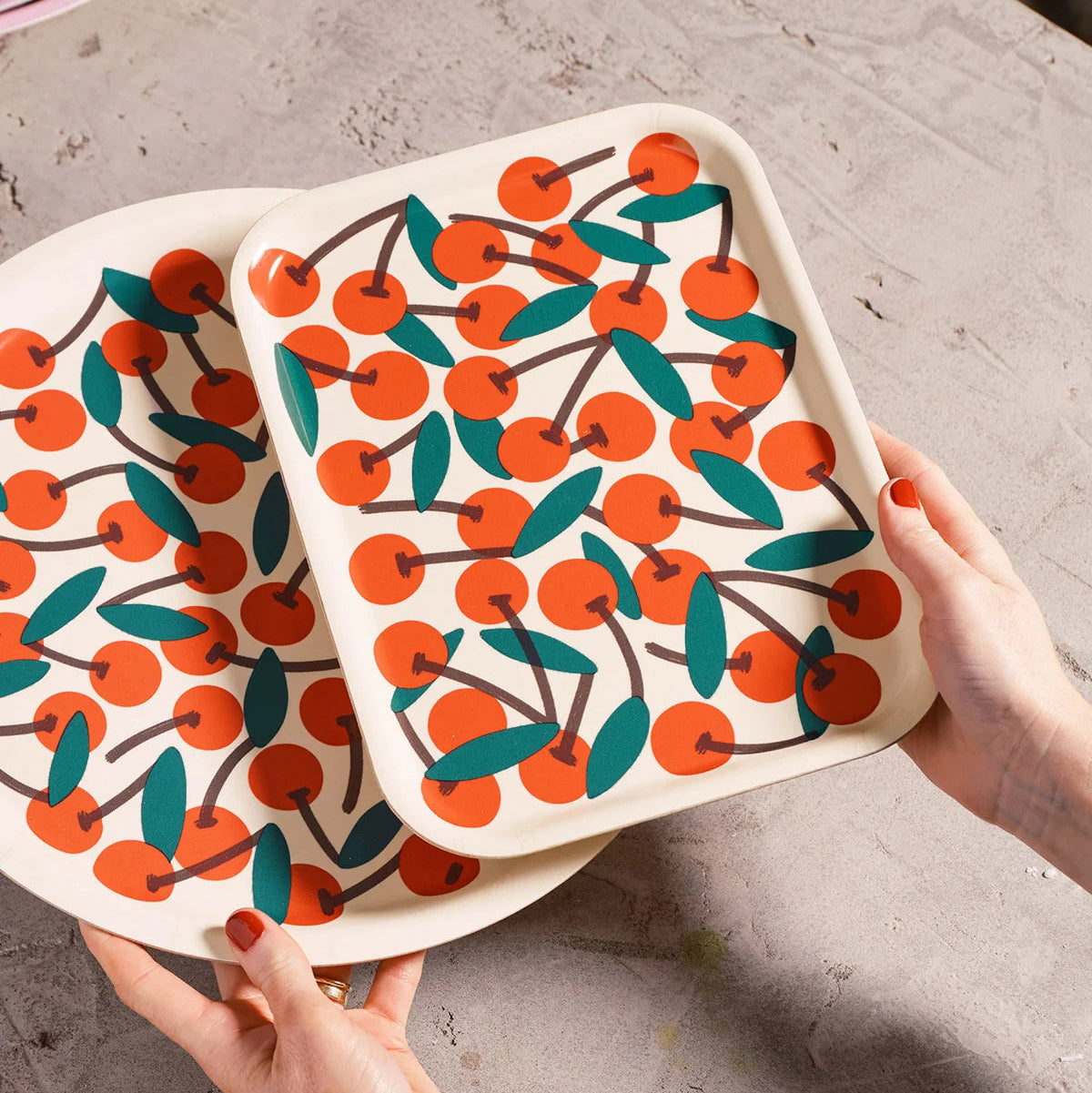 Two square ceramic trays with rounded corners featuring a vibrant orange cherry and green leaf pattern on a cream background, held by hands over a textured gray stone surface.
