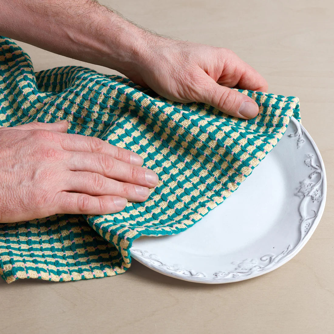 Person using a green and yellow checkered cloth to wipe a white plate on a beige surface