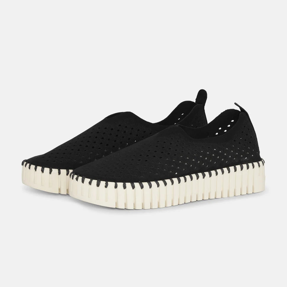 Black perforated slip-on sneakers with a cream-colored, textured sole. The shoes have a casual, comfortable style.