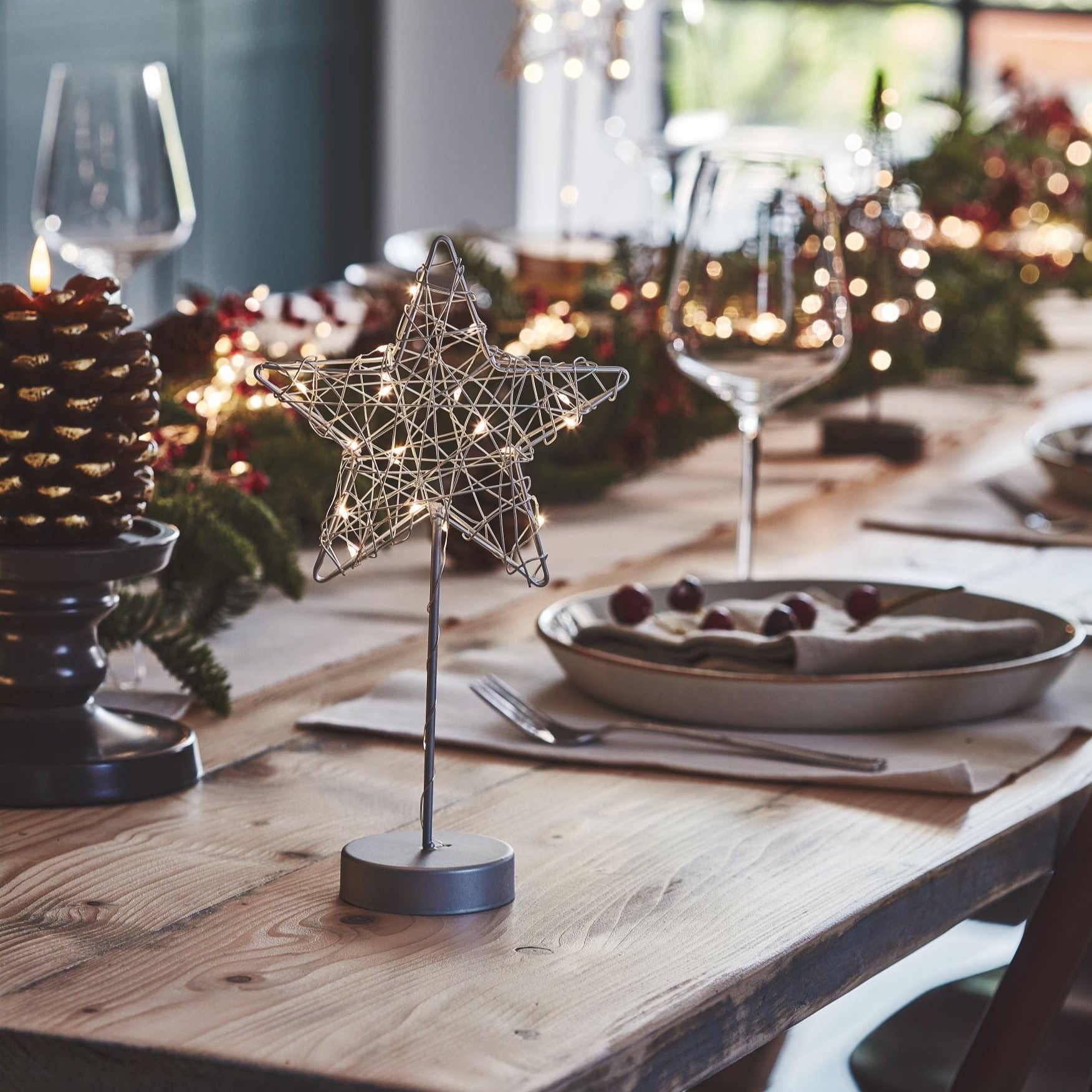 Silver wire star-shaped LED light on a wooden table.  The star is illuminated and features a small, round base.