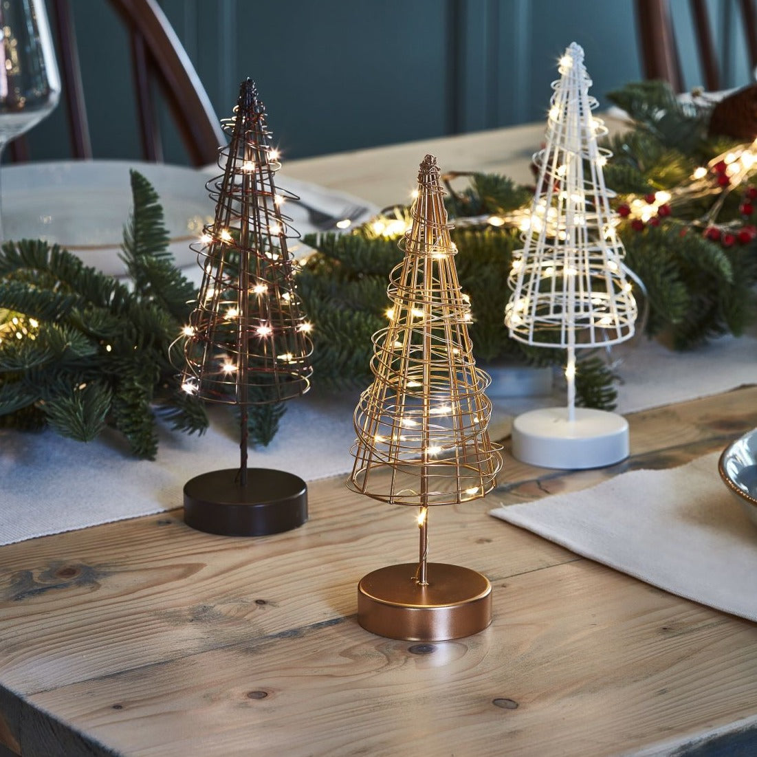 Three miniature wire Christmas trees with warm white LED lights.  Trees are in copper, dark bronze, and white metal.