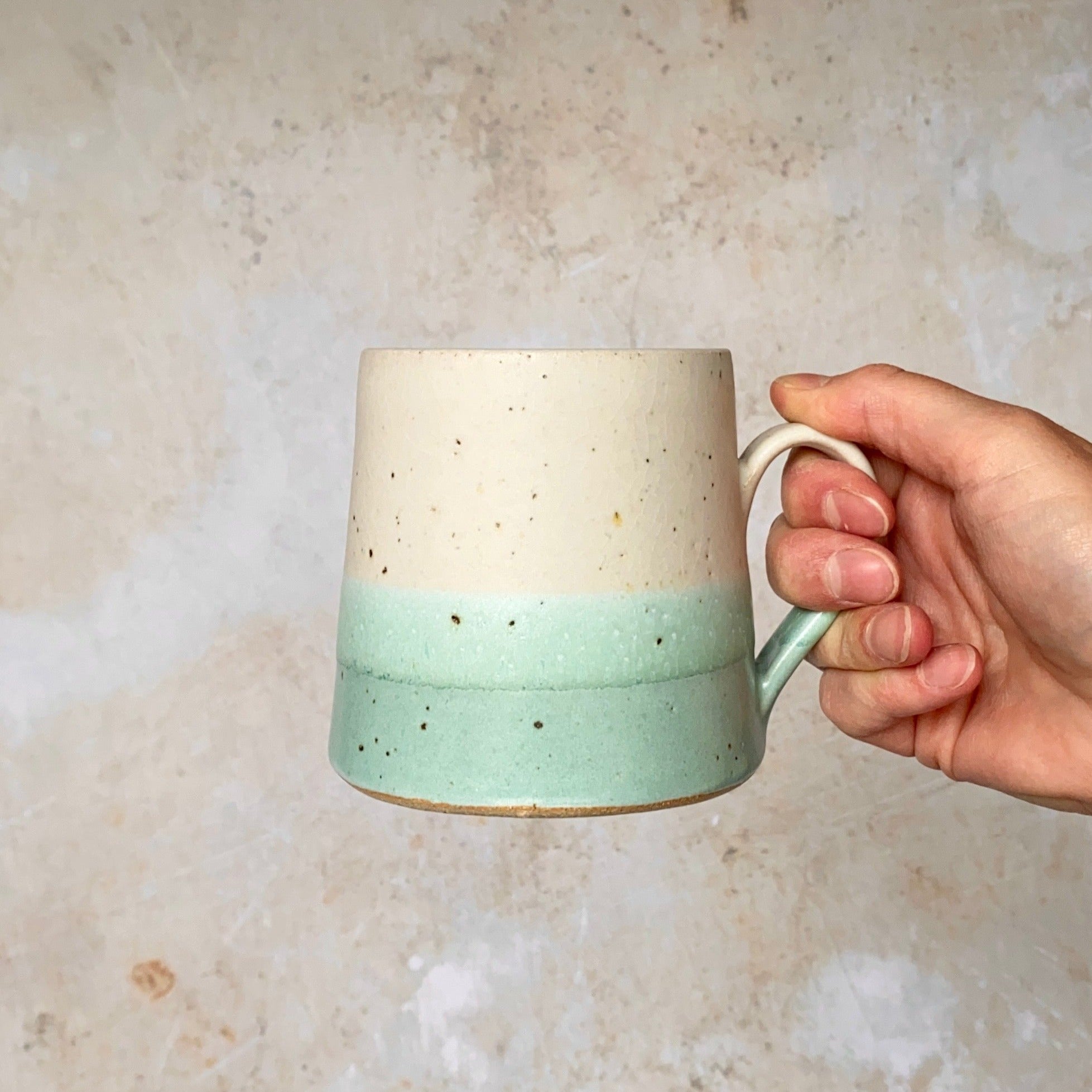 A speckled off-white and light teal ceramic mug with a handle, held in a hand.