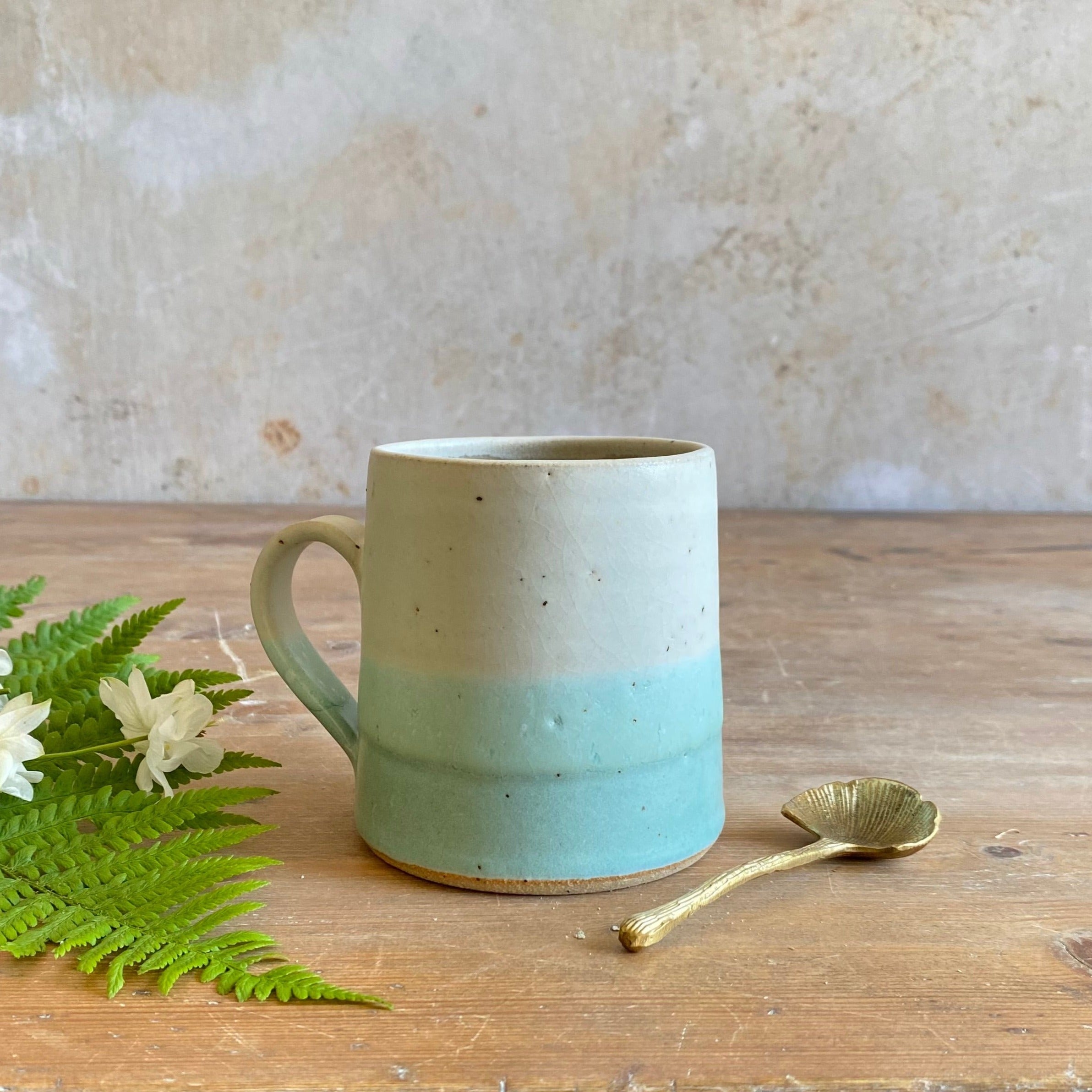 Stoneware mug with speckled off-white top and light teal bottom.  The mug has a slightly curved handle.