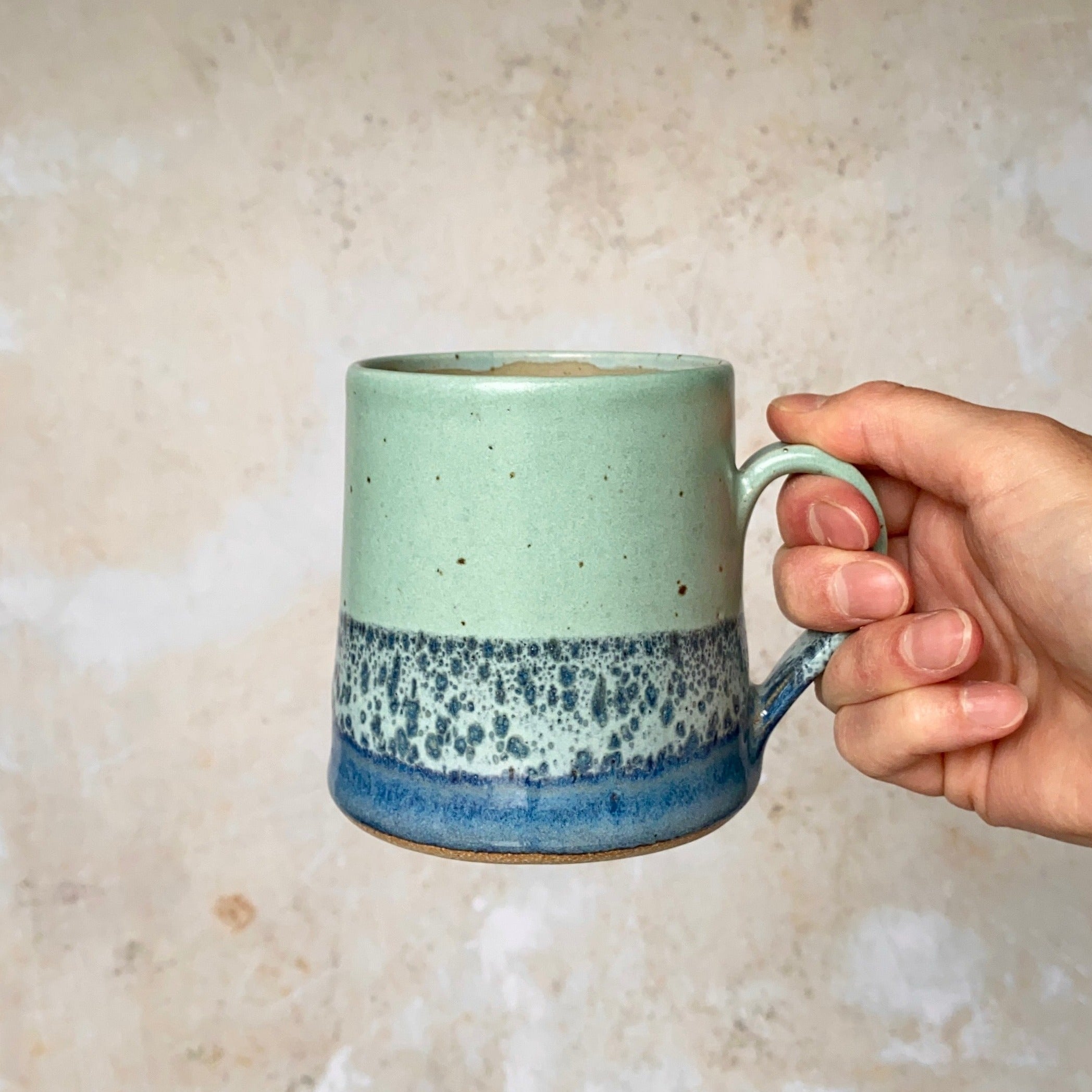 Hand holding a light green and blue speckled ceramic mug with a handle.