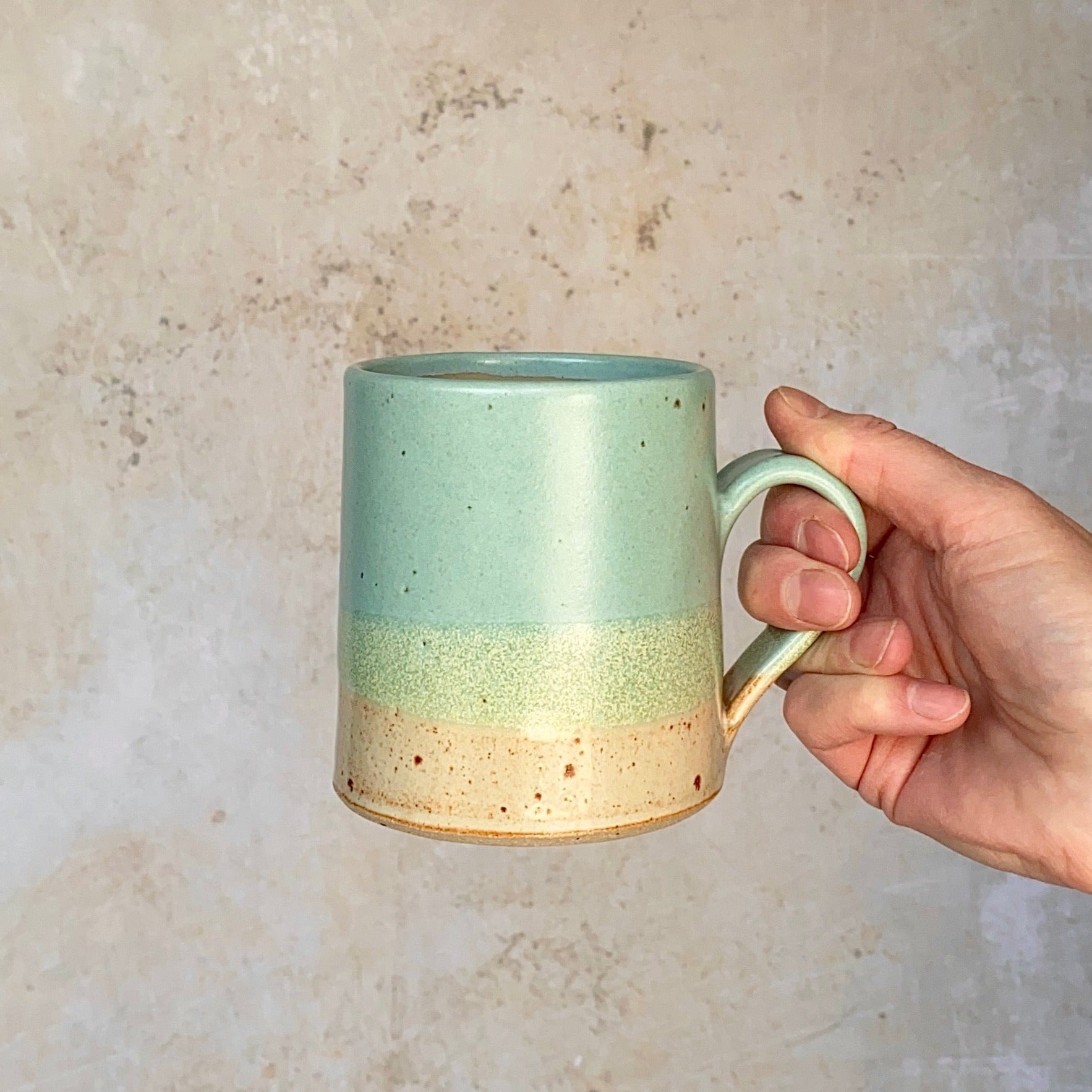 Hand holding a light teal and beige speckled ceramic mug.